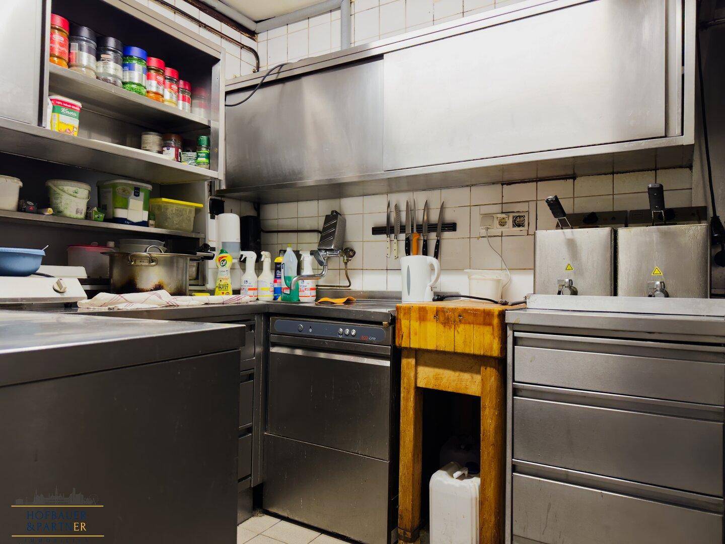 Professionelle Gastroküche mit Edelstahloberflächen, Spülmaschine und Regalen für Vorräte, funktional ausgestattet.