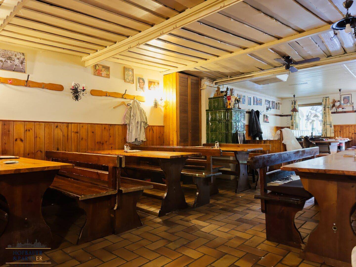 Geräumiger Gastraum mit vielen Sitzgelegenheiten, Holzelementen und einem markanten Kachelofen im Zentrum.