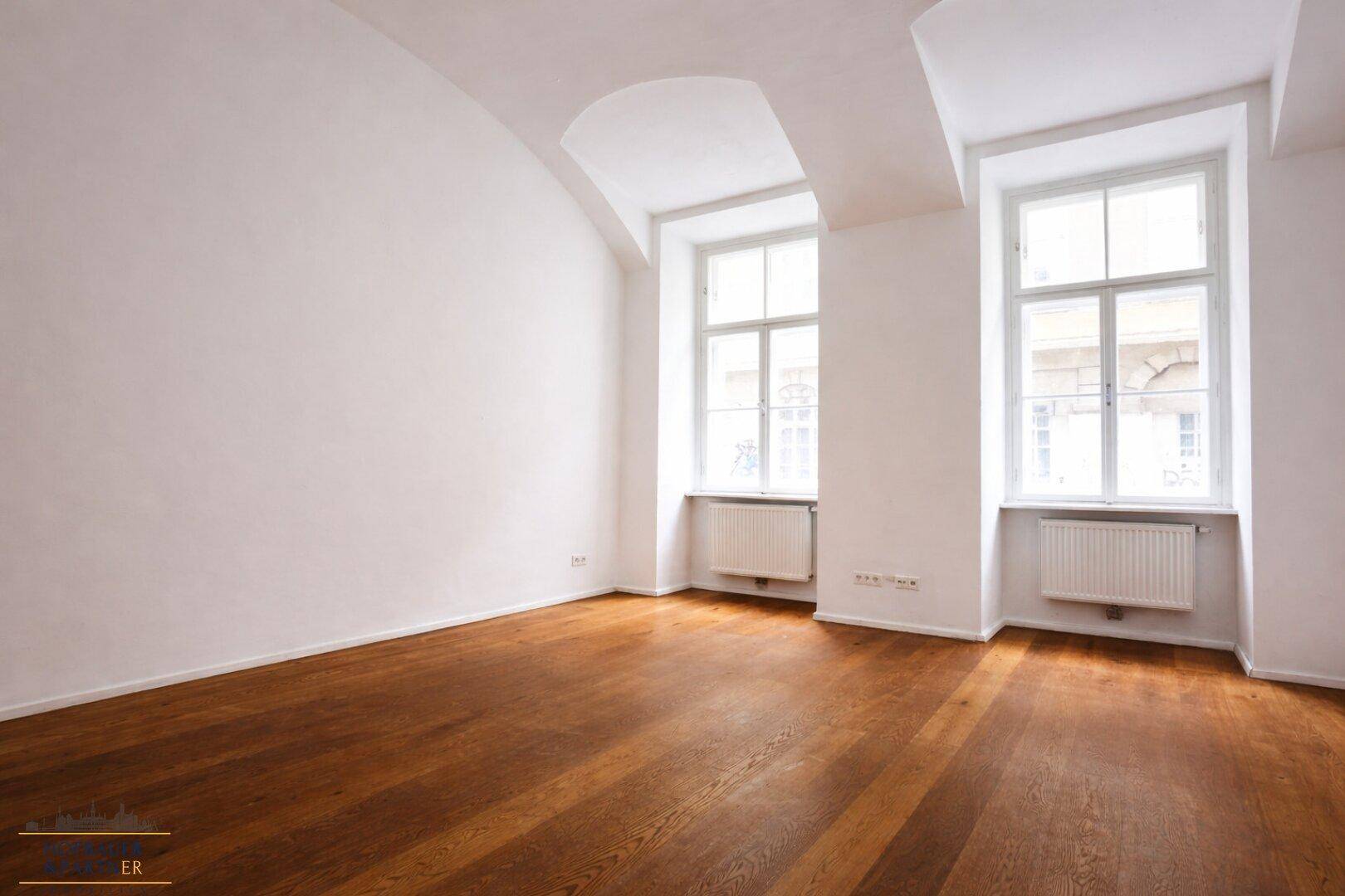 Geräumiger Raum mit Holzboden, weißen Wänden und zwei hohen Fenstern, die viel Tageslicht bieten.