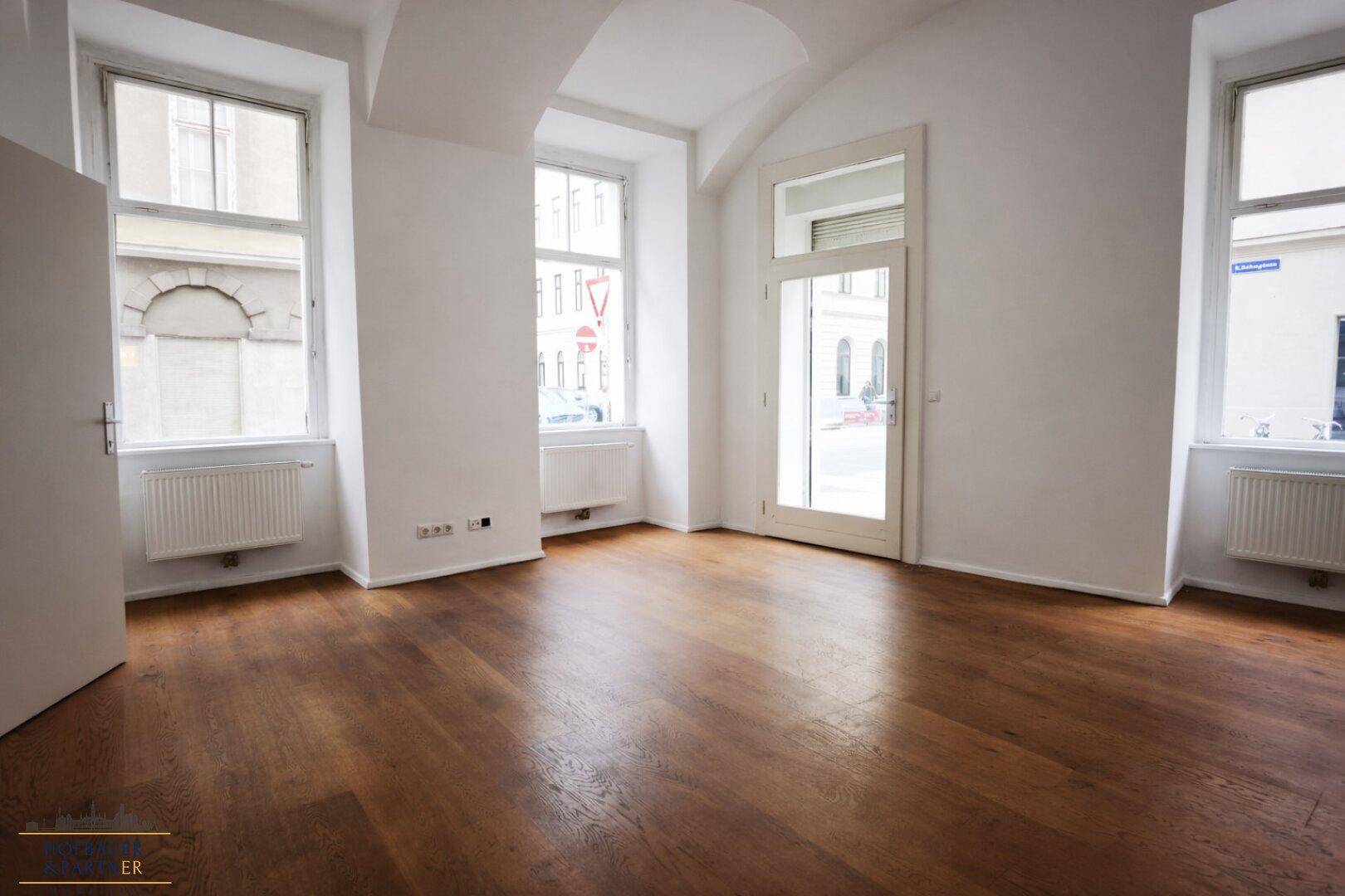 Heller Raum mit Holzboden, großen Fenstern und einer Glastür, die viel Licht hereinlässt.
