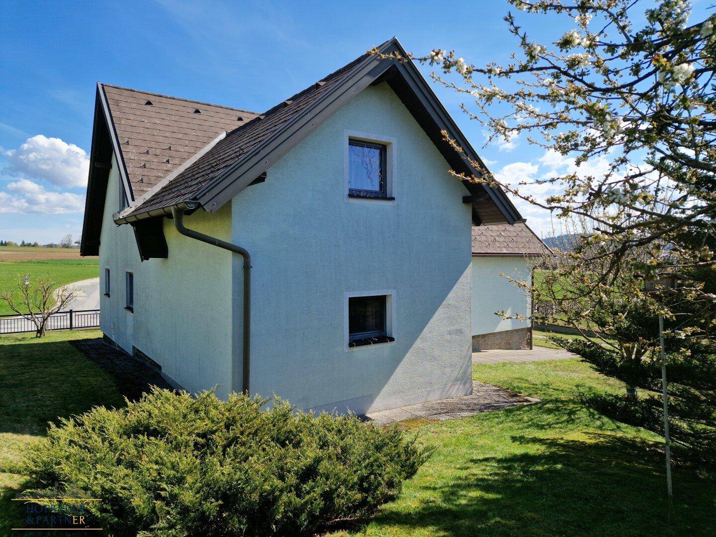 Seitenansicht des Hauses mit weißer Fassade, Dachschräge und grüner Bepflanzung.