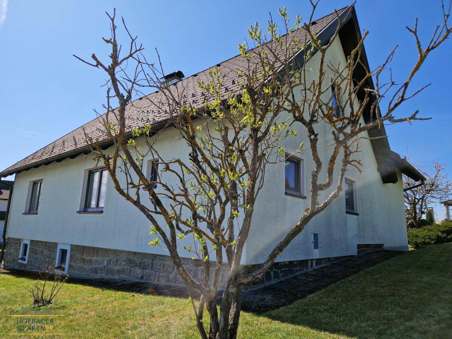 Hausansicht mit weißer Fassade, Satteldach und blühendem Baum im Vorgarten.