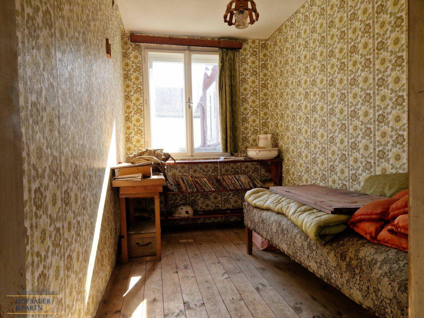 Schlafzimmer mit gemusterter Tapete, Holzdielen und einem Fenster.