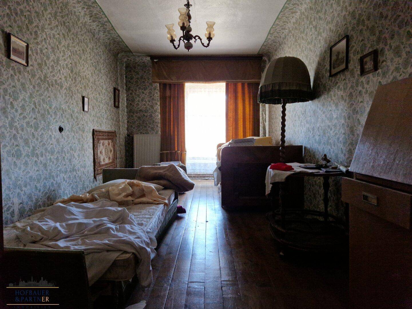 Schlafzimmer mit dunklen Holzdielen, gemusterter Tapete und einem Kronleuchter.