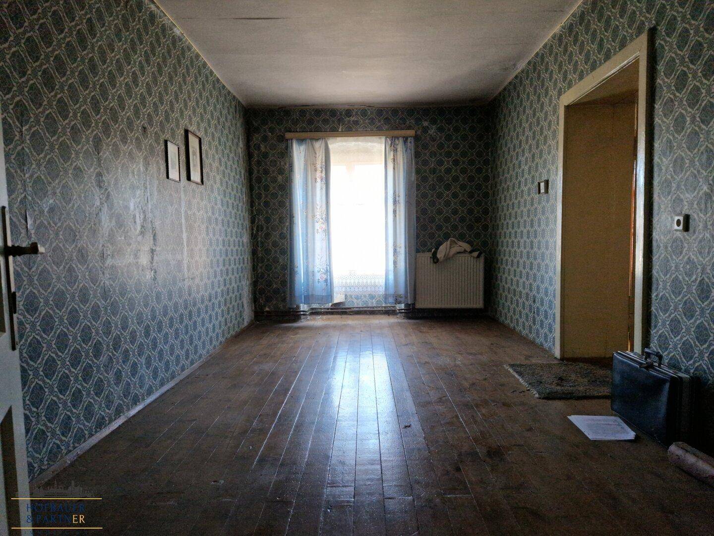 Leerer Raum mit gemusterter Tapete, Holzdielen und einem Fenster.