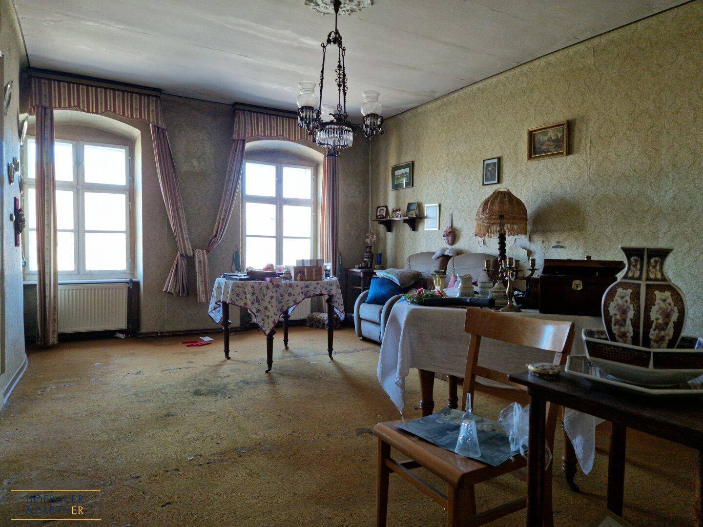 Geräumiges Wohnzimmer mit mehreren Fenstern, Teppichboden und traditionellen Möbeln.