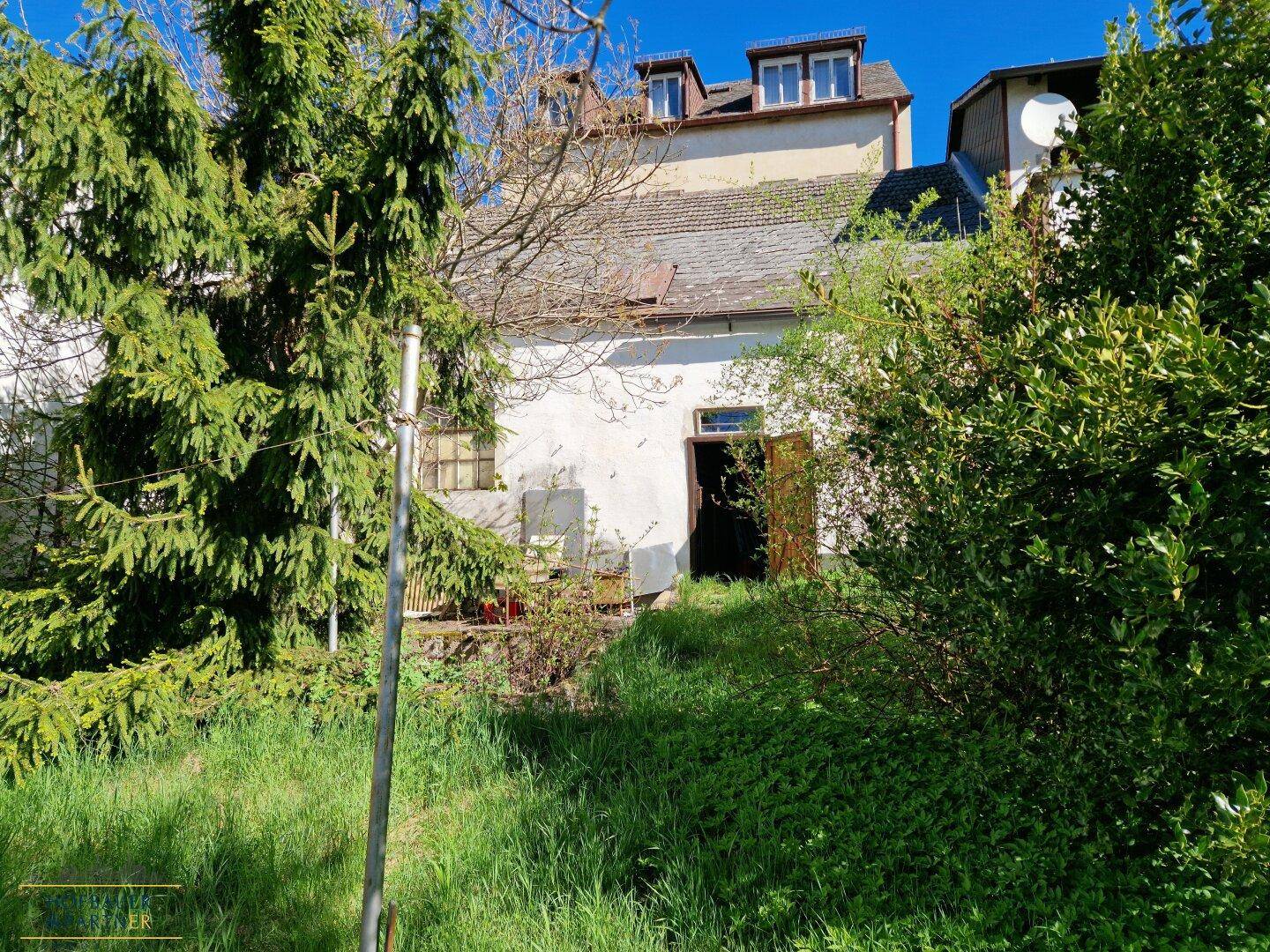 Gartenbereich mit Bäumen, Sträuchern und einem alten Nebengebäude.