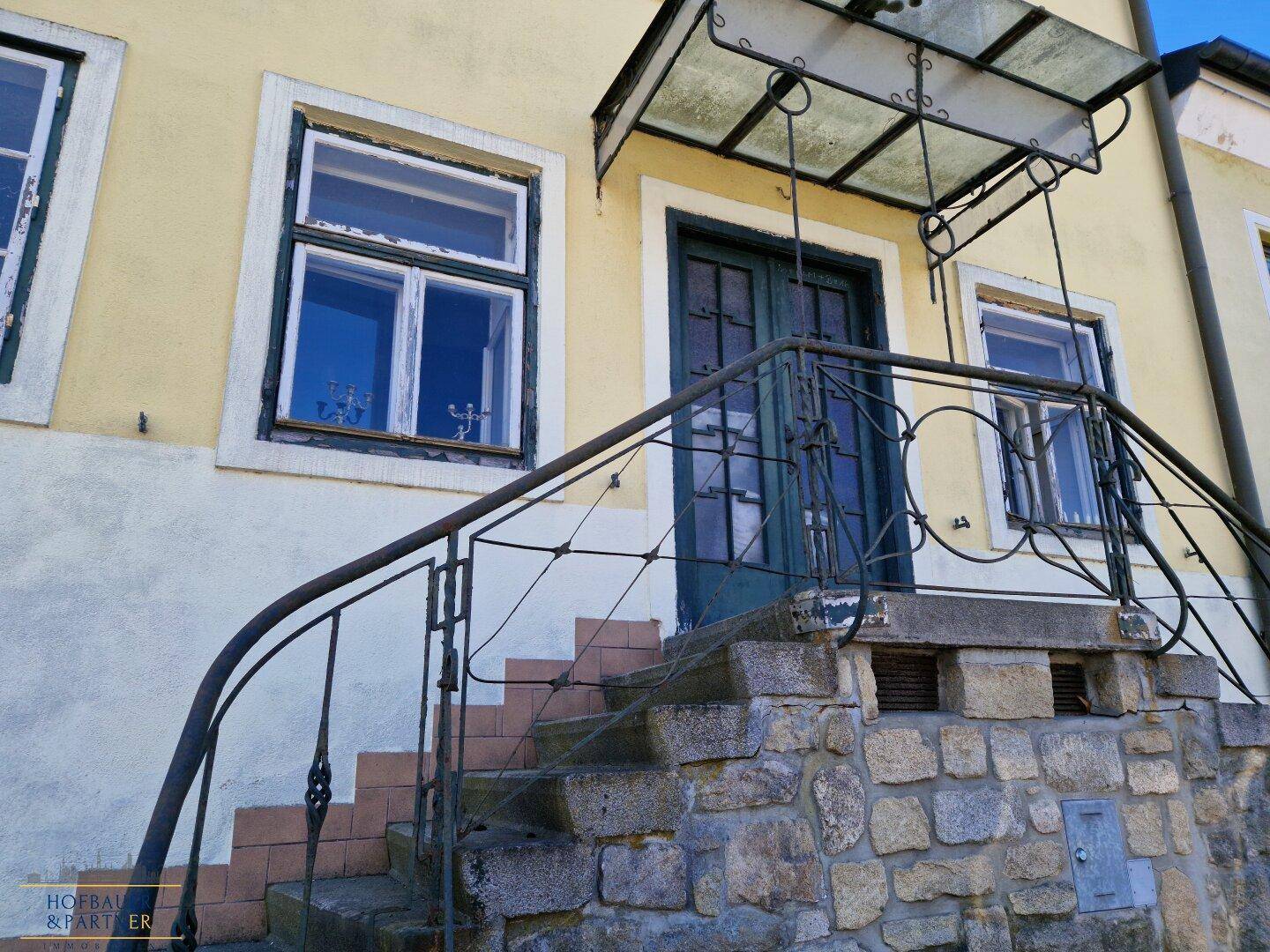 Eingangsbereich des Gebäudes mit Steintreppe, Geländer und Vordach.