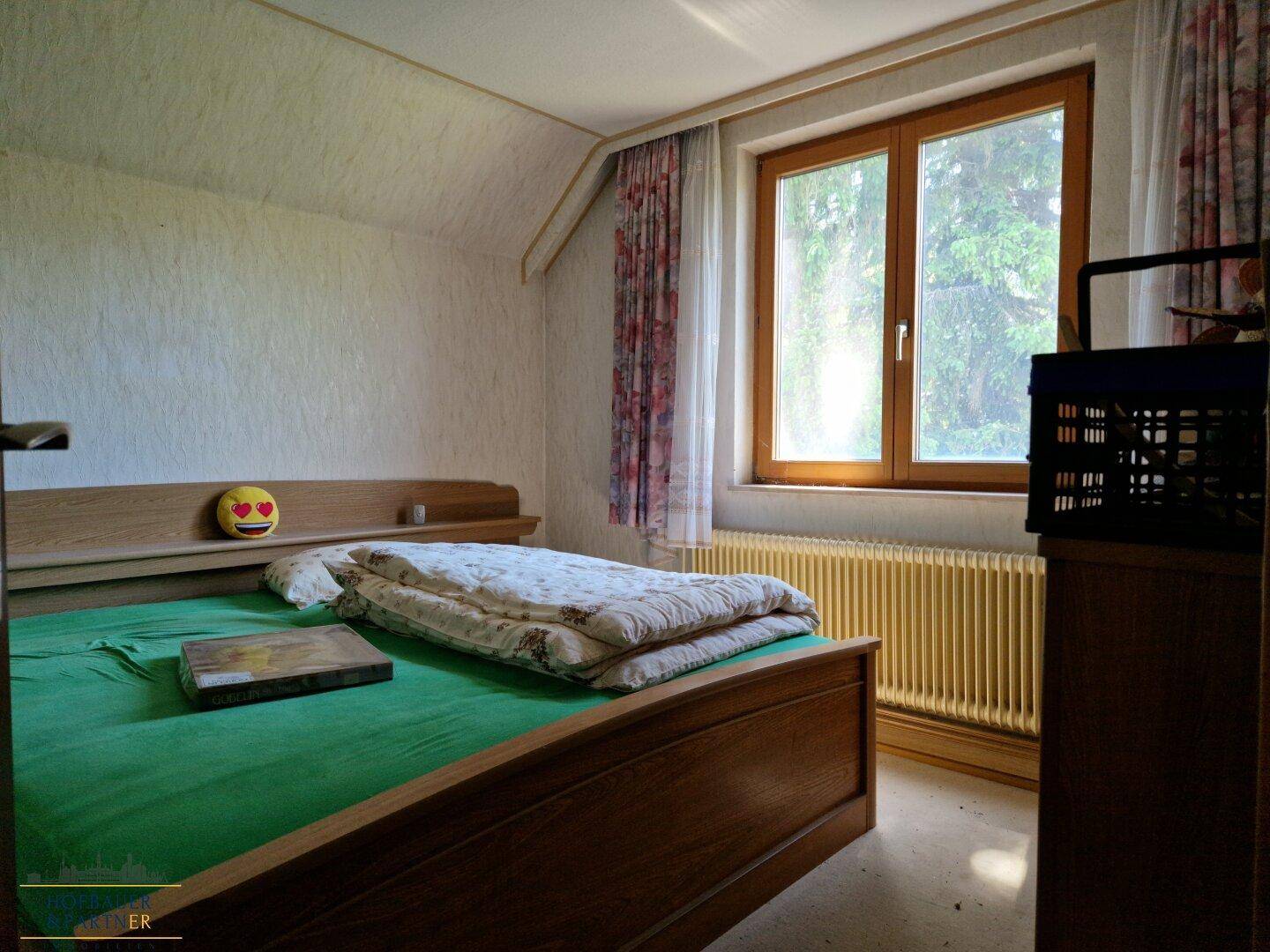 Schlafzimmer mit Holzbett, Fenster und Heizkörper, umgeben von einfacher Einrichtung.