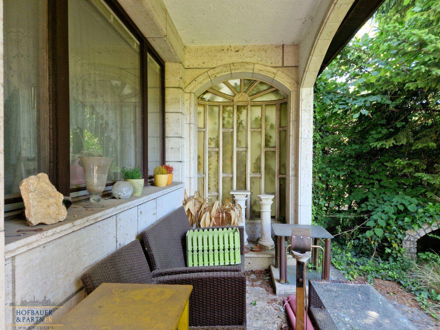 Überdachte Terrasse mit Sitzgelegenheiten und dekorativen Elementen, umgeben von viel Grün.