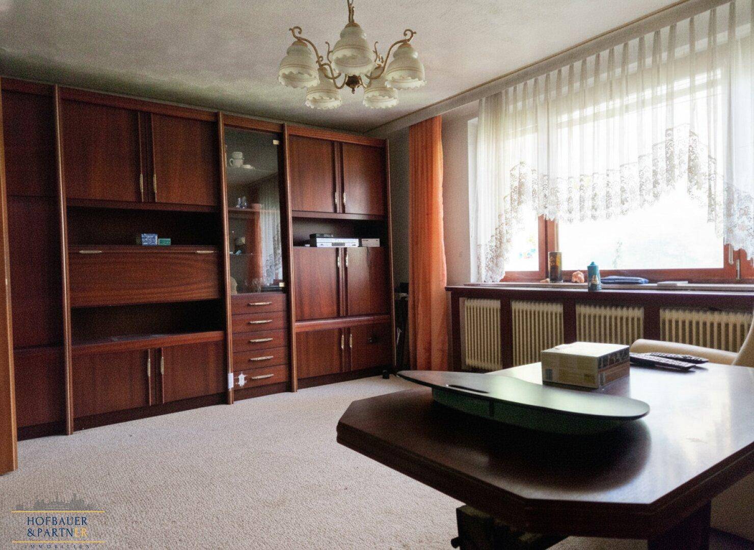 Geräumiges Wohnzimmer mit Teppichboden, großen Fenstern und traditionellen Holzmöbeln.