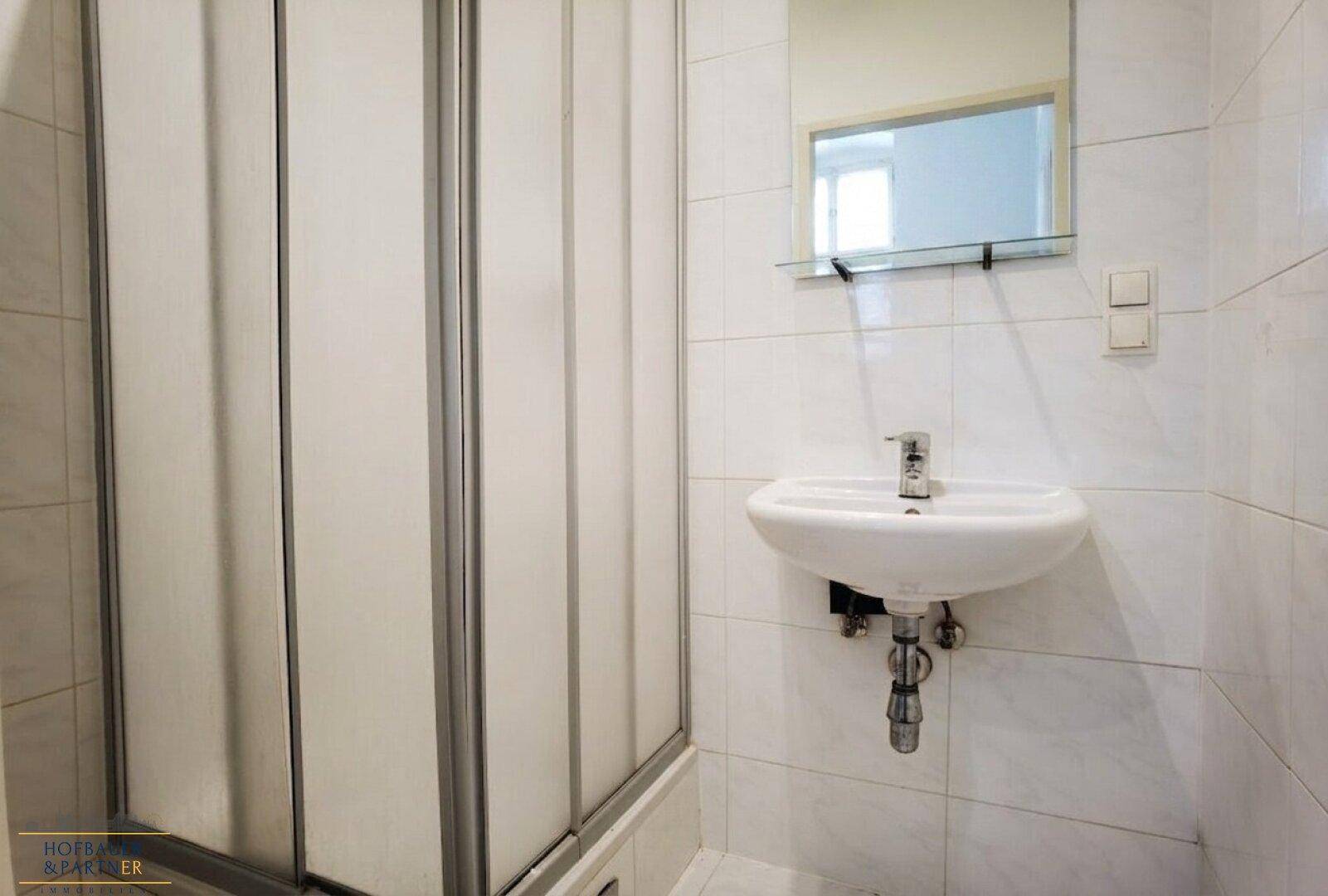 Helles Badezimmer mit weiß gefliesten Wänden, einer Duschkabine und einem wandmontierten Waschbecken mit Spiegel.