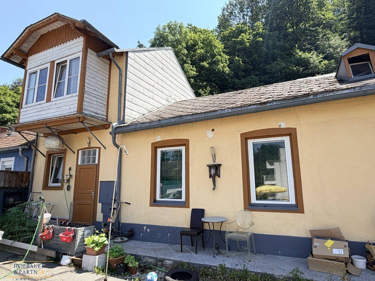 Detailansicht der gelben Fassade mit Fenstern, Eingangstür und einem kleinen Bereich für Sitzgelegenheiten.