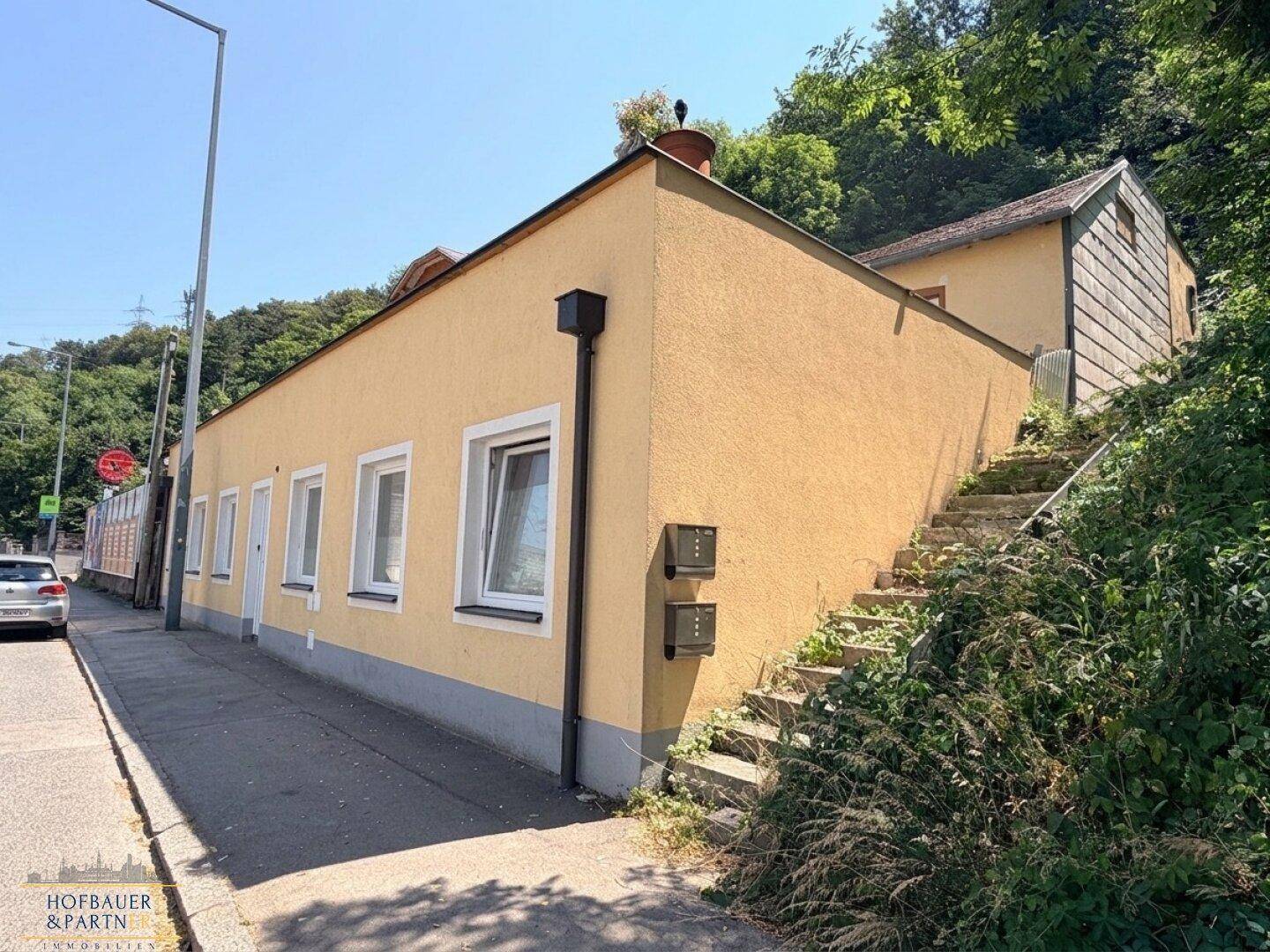 Seitenansicht des Gebäudes mit gelber Fassade, Fenstern und einer Außentreppe zum Hang.