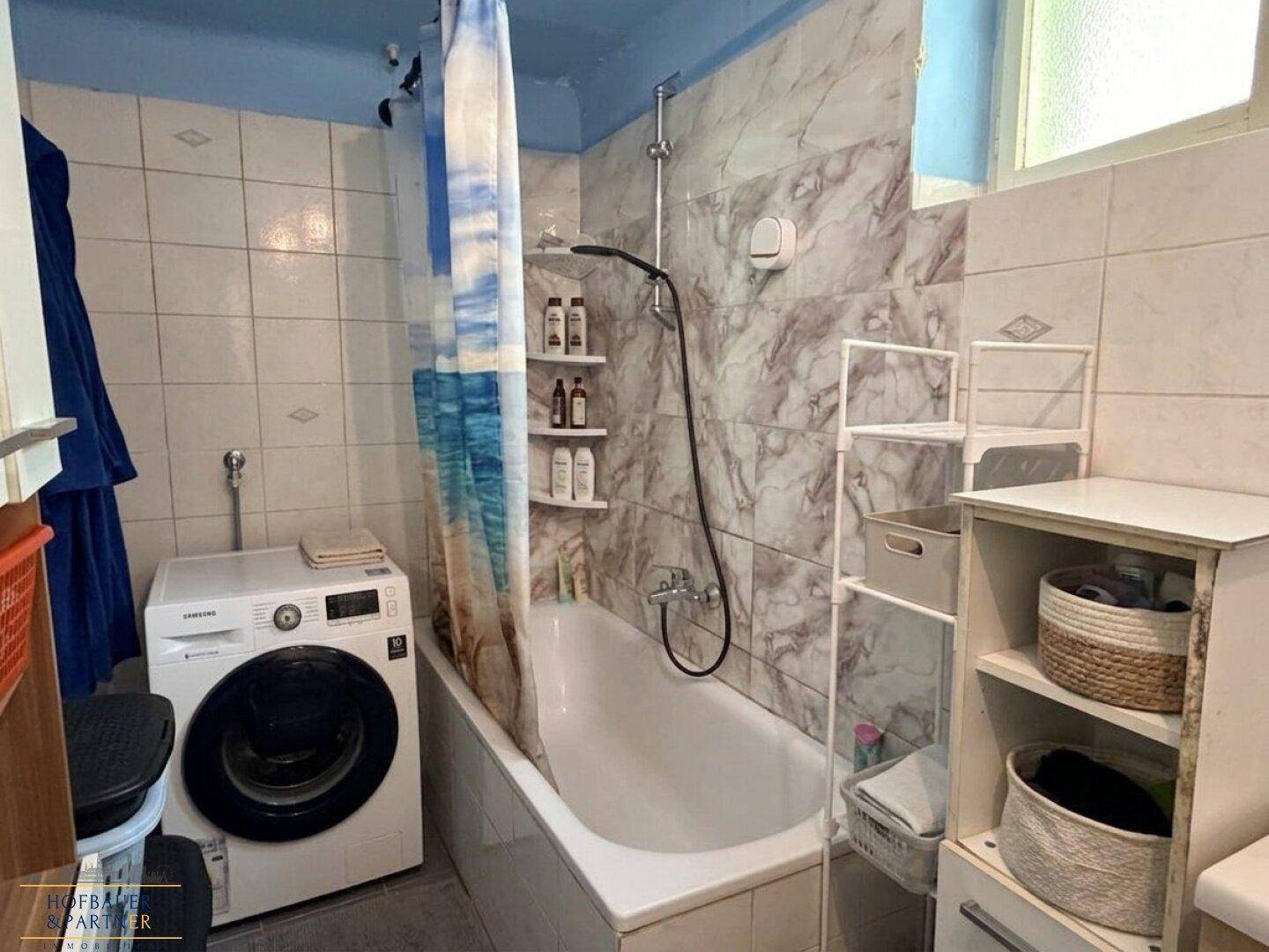 Badezimmer mit Badewanne, Duschvorhang, Waschmaschine und Fliesen an Wänden und Boden.