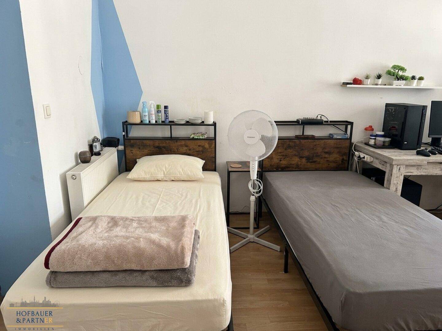 Helles Schlafzimmer mit zwei Einzelbetten, Holzfußboden und einem Ventilator.