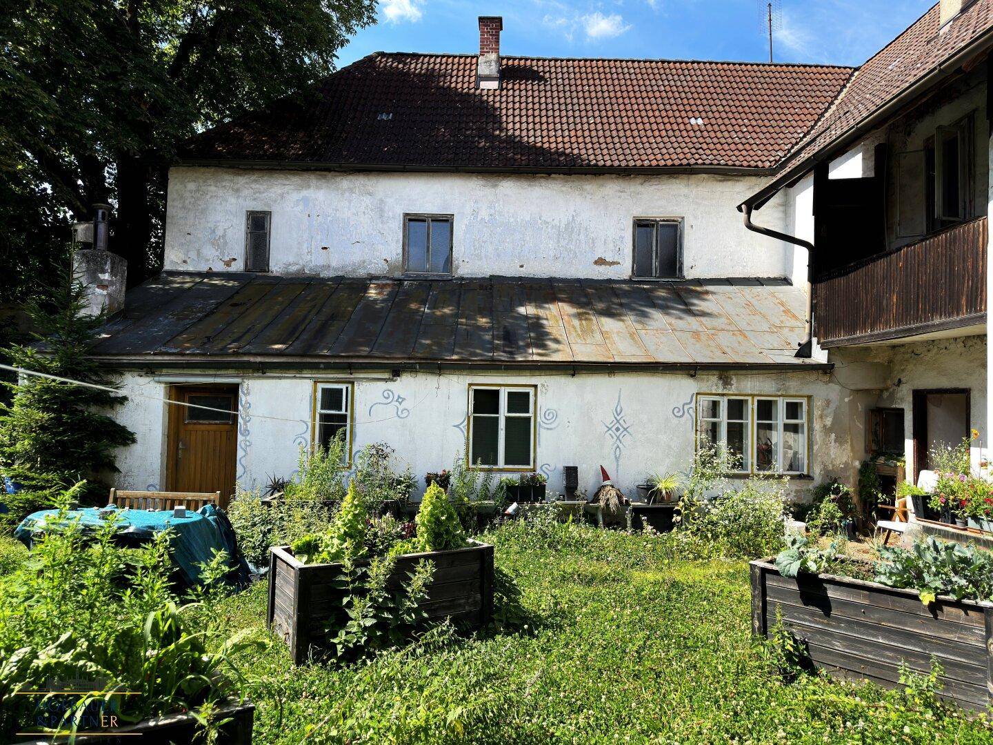 Gartenbereich mit rustikalen Hochbeeten, einer kleinen Sitzecke und einem Gebäude mit dekorativer Fassadenmalerei.