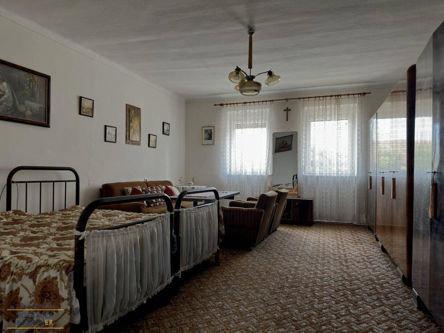 Gemütliches Schlafzimmer mit Doppelbett, Sofa, Teppichboden und viel natürlichem Licht durch zwei Fenster.