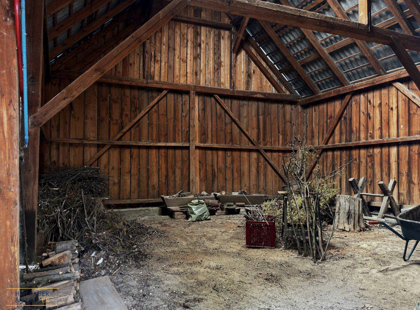 Geräumige Scheune mit sichtbaren Holzbalken, Holzwänden und gelagertem Brennholz und Ästen.