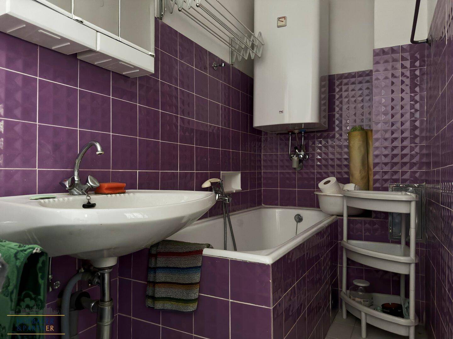 Badezimmer mit auffälligen lila Fliesen, Waschbecken, Badewanne und einem Warmwasserboiler.