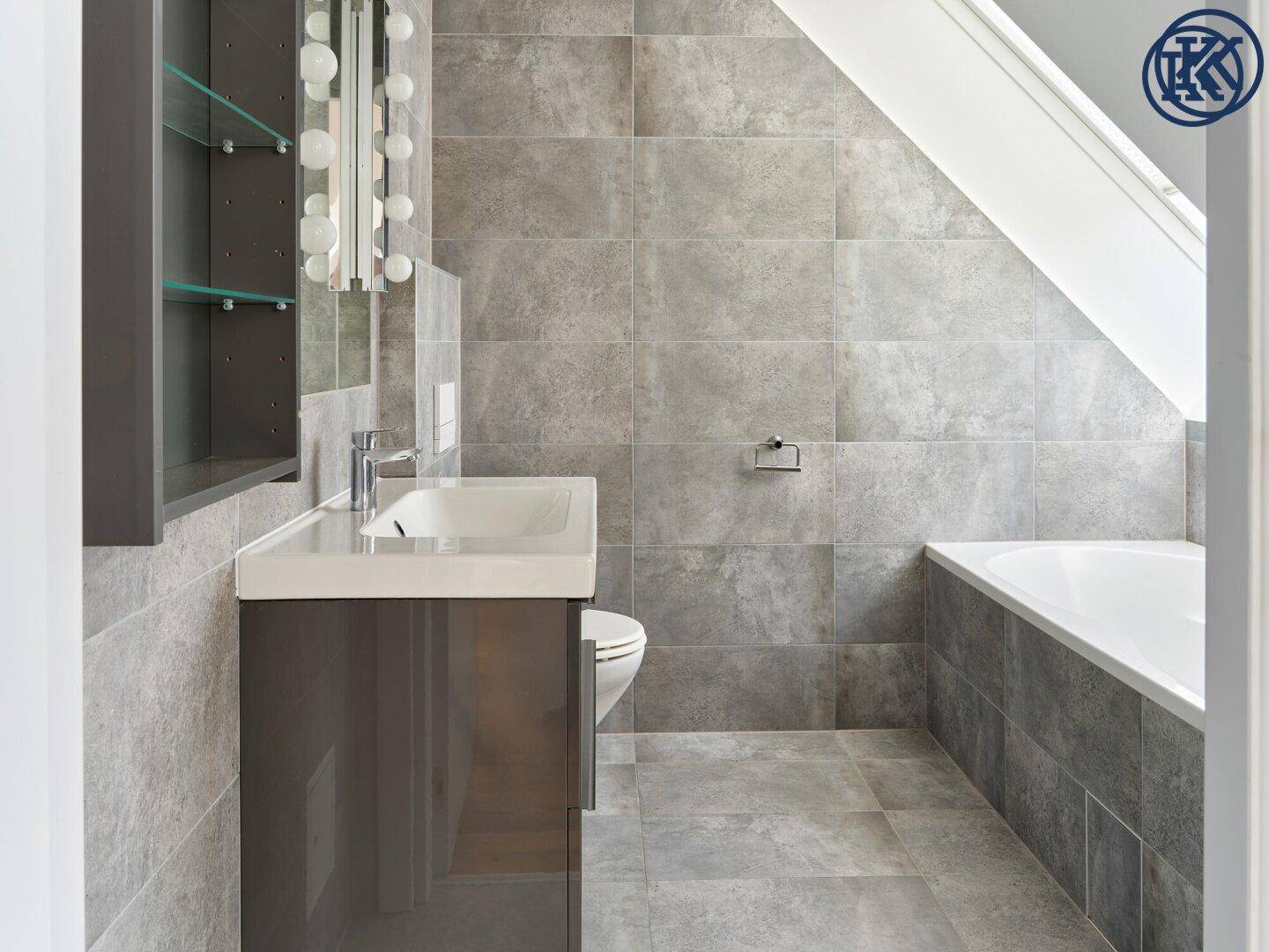 Modernes Badezimmer mit grauen Fliesen, Waschtisch, Toilette und Badewanne unter einem Dachfenster.