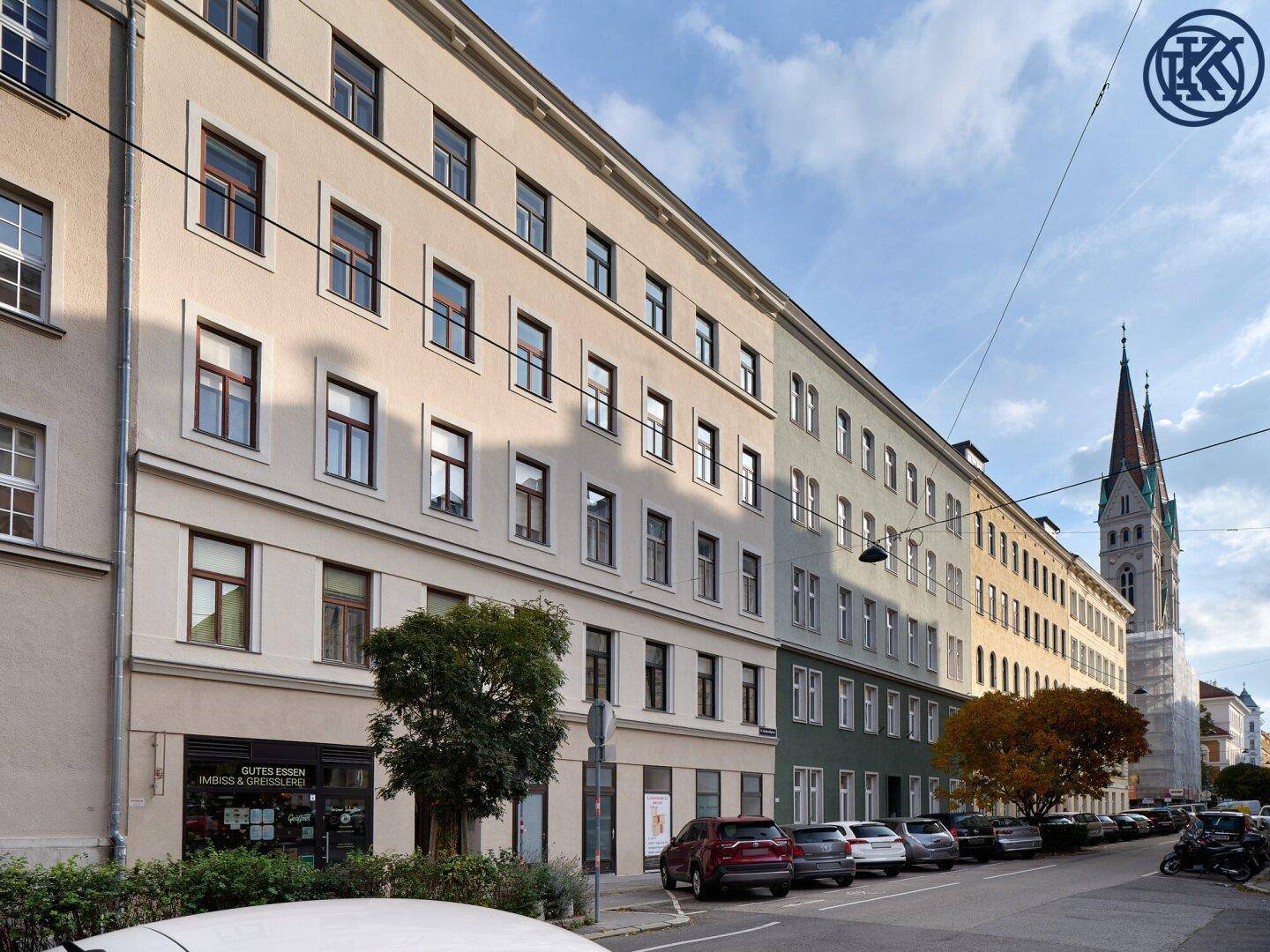 Ansicht einer belebten Stadtstraße mit geparkten Autos und einem hohen Kirchturm im Hintergrund.