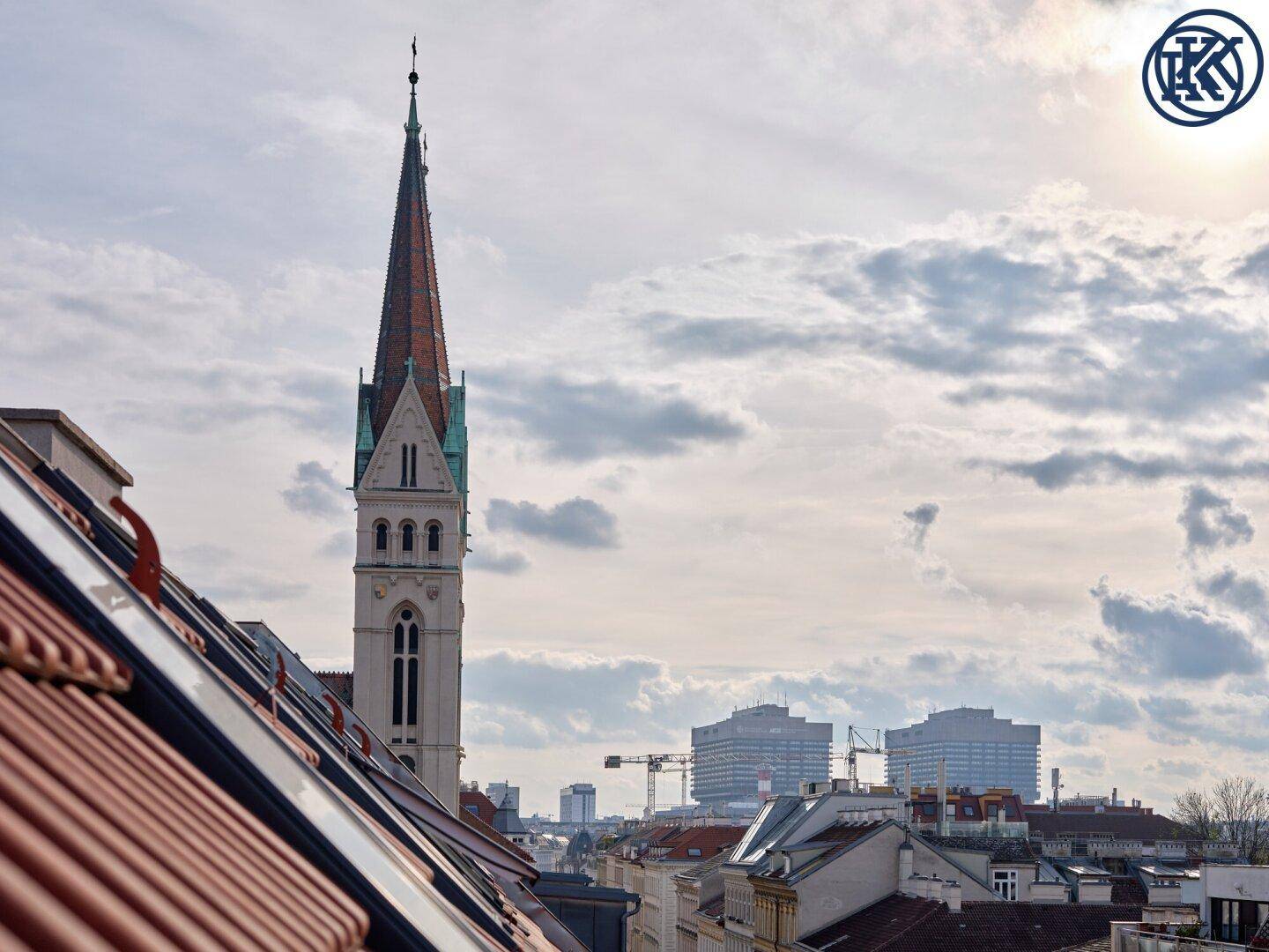Blick über die Dächer der Stadt mit einem hohen Kirchturm und modernen Gebäuden im Hintergrund.