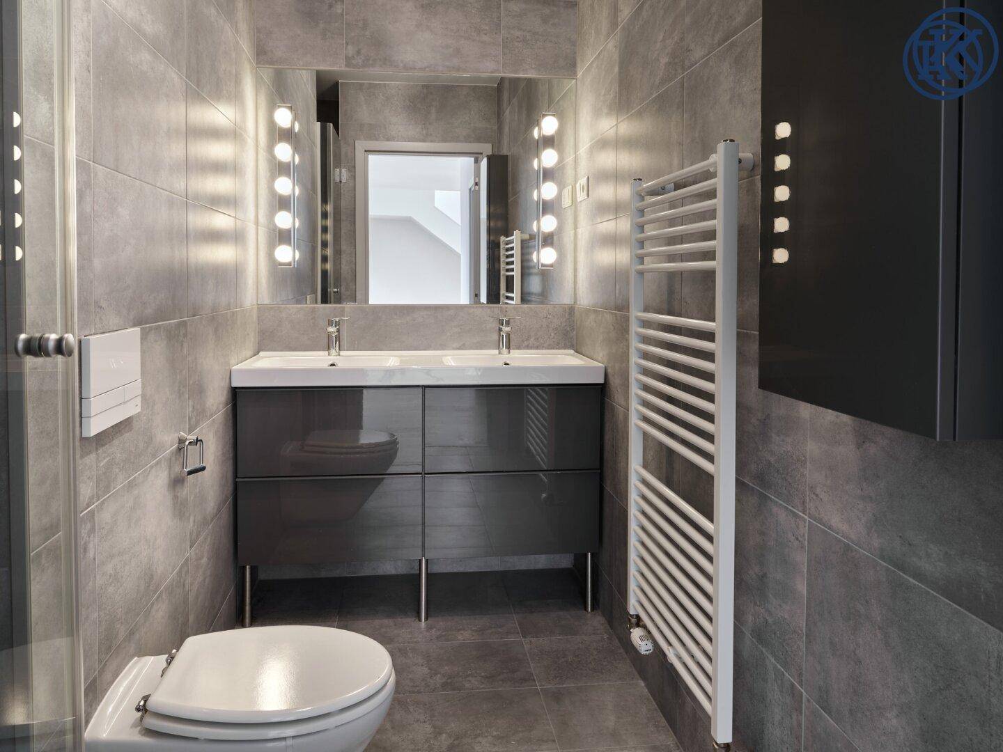 Modernes Badezimmer mit grauen Fliesen, Doppelwaschbecken, WC und beheiztem Handtuchhalter.