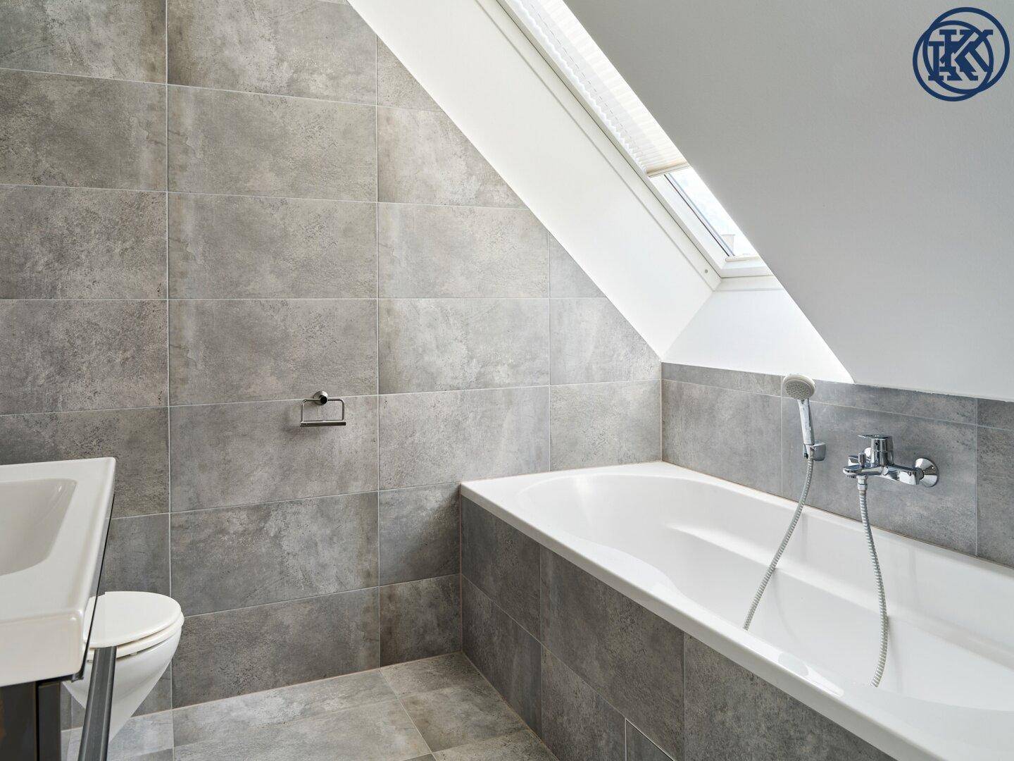 Helles Badezimmer mit grauen Fliesen, Badewanne und Dachfenster, das viel Tageslicht spendet.