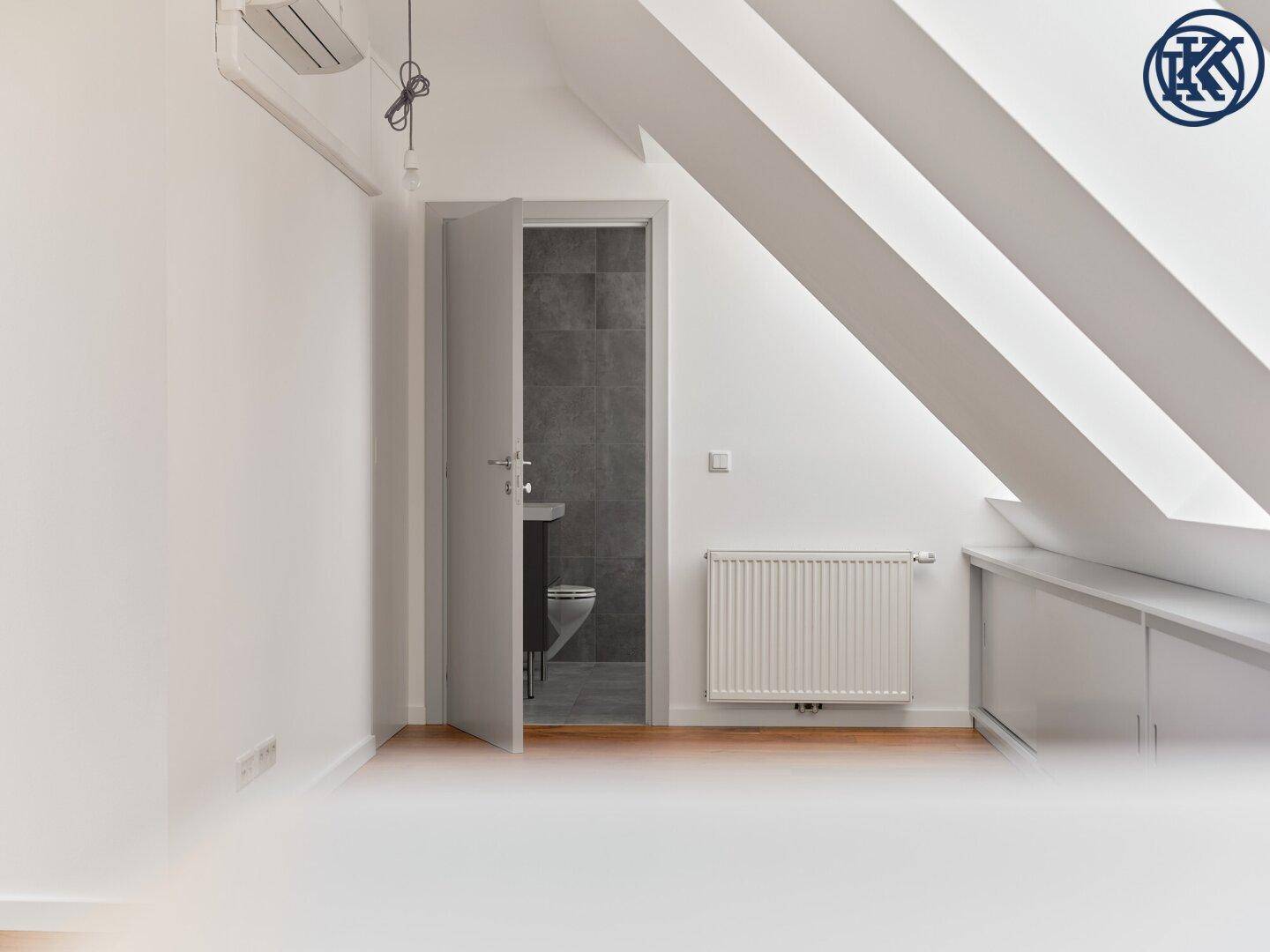 Leeres Dachzimmer mit Holzboden, Heizkörper und Zugang zu einem modernen Badezimmer.