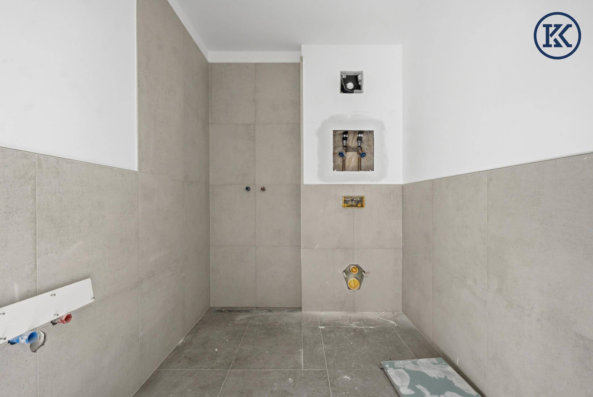 Badezimmer im Rohbau mit hellen Fliesen und vorbereiteten Anschlüssen für Sanitäranlagen.