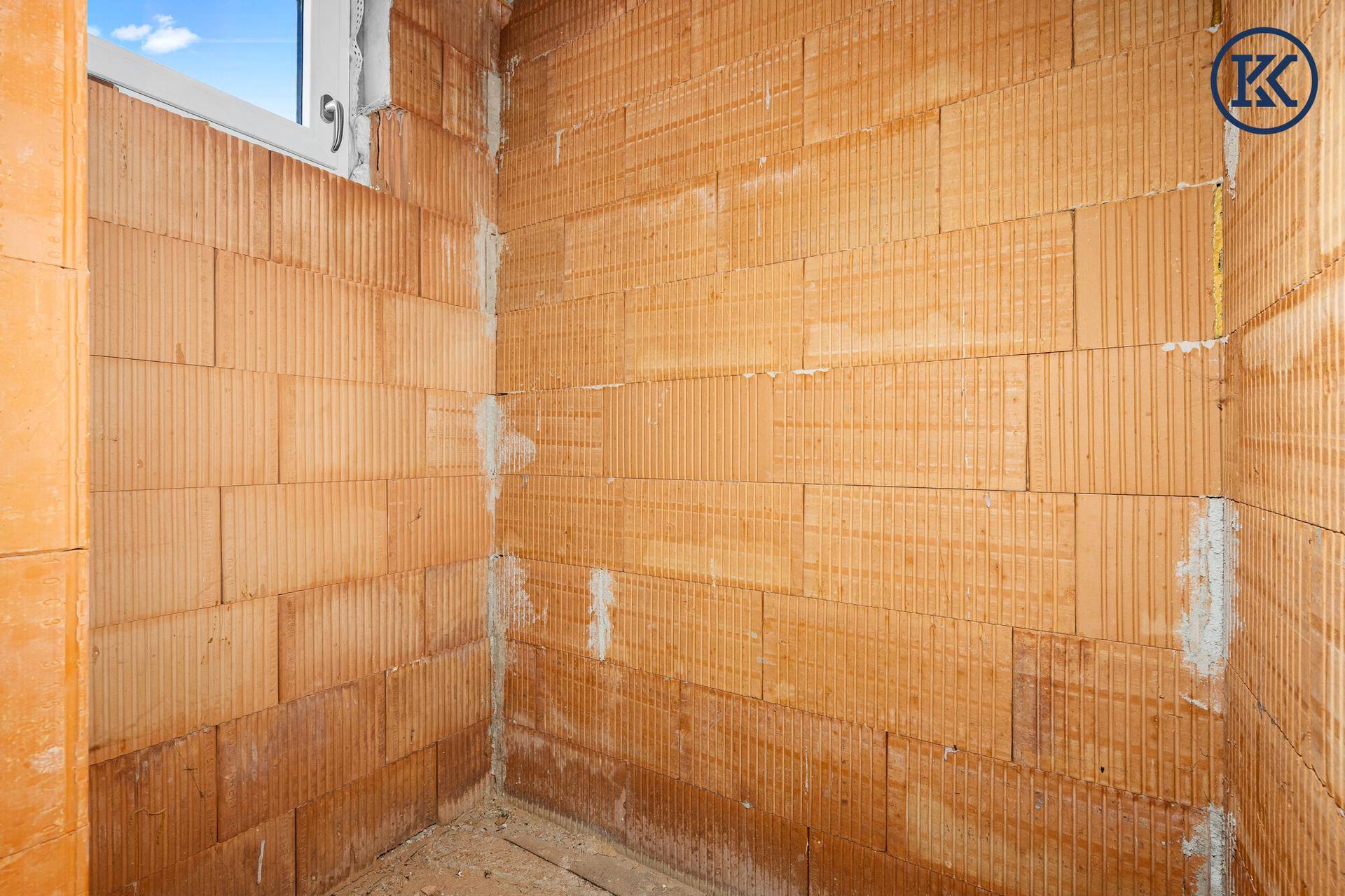 Kleiner Raum im Rohbau mit Ziegelwänden und einem Fenster, das Tageslicht hereinlässt.