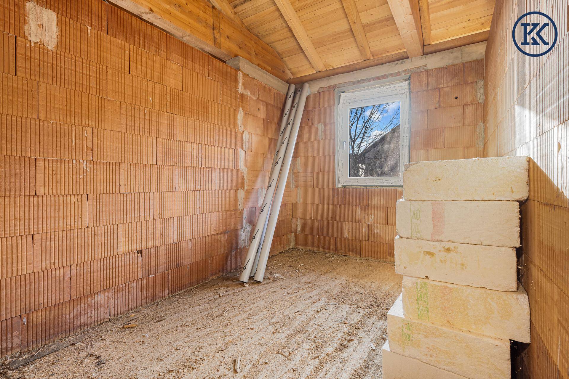 Rohbau-Raum mit Ziegelwänden, Fenster und gestapelten Baumaterialien, viel Tageslicht.