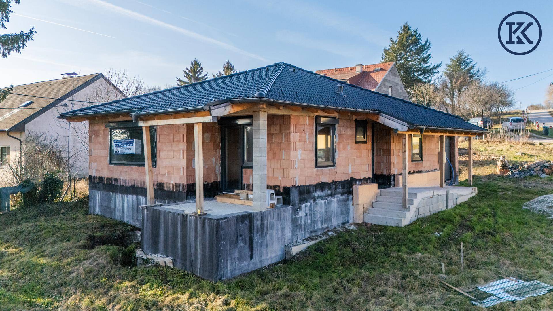 Rohbauhaus mit dunklem Dach und Ziegelwänden, umgeben von einer grünen Wiese und Bäumen.