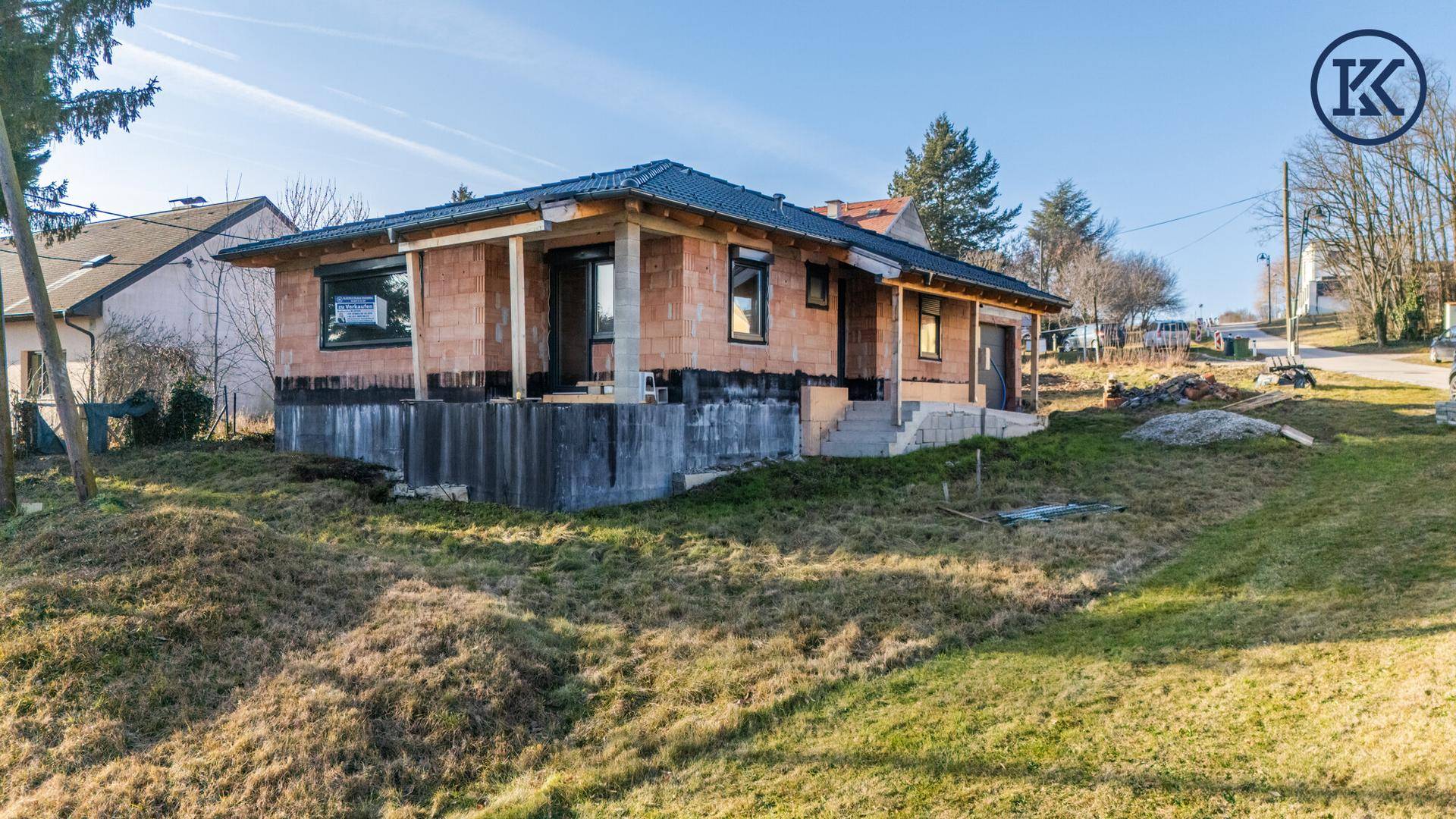 Rohbauhaus mit dunklem Dach und Ziegelwänden, umgeben von einer grünen Wiese und Bäumen.