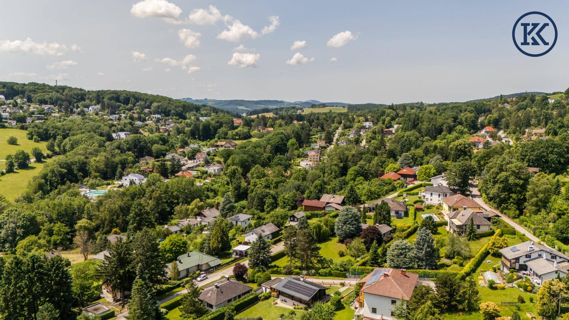 Weitläufige Luftaufnahme einer idyllischen Wohngegend mit viel Grün und Hügeln.