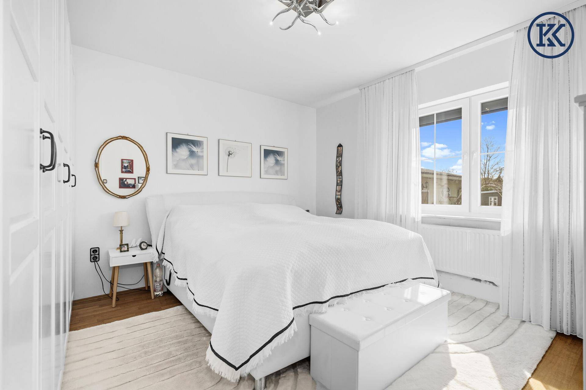 Ruhiges Schlafzimmer mit weißem Doppelbett, Parkettboden und großem Fenster.