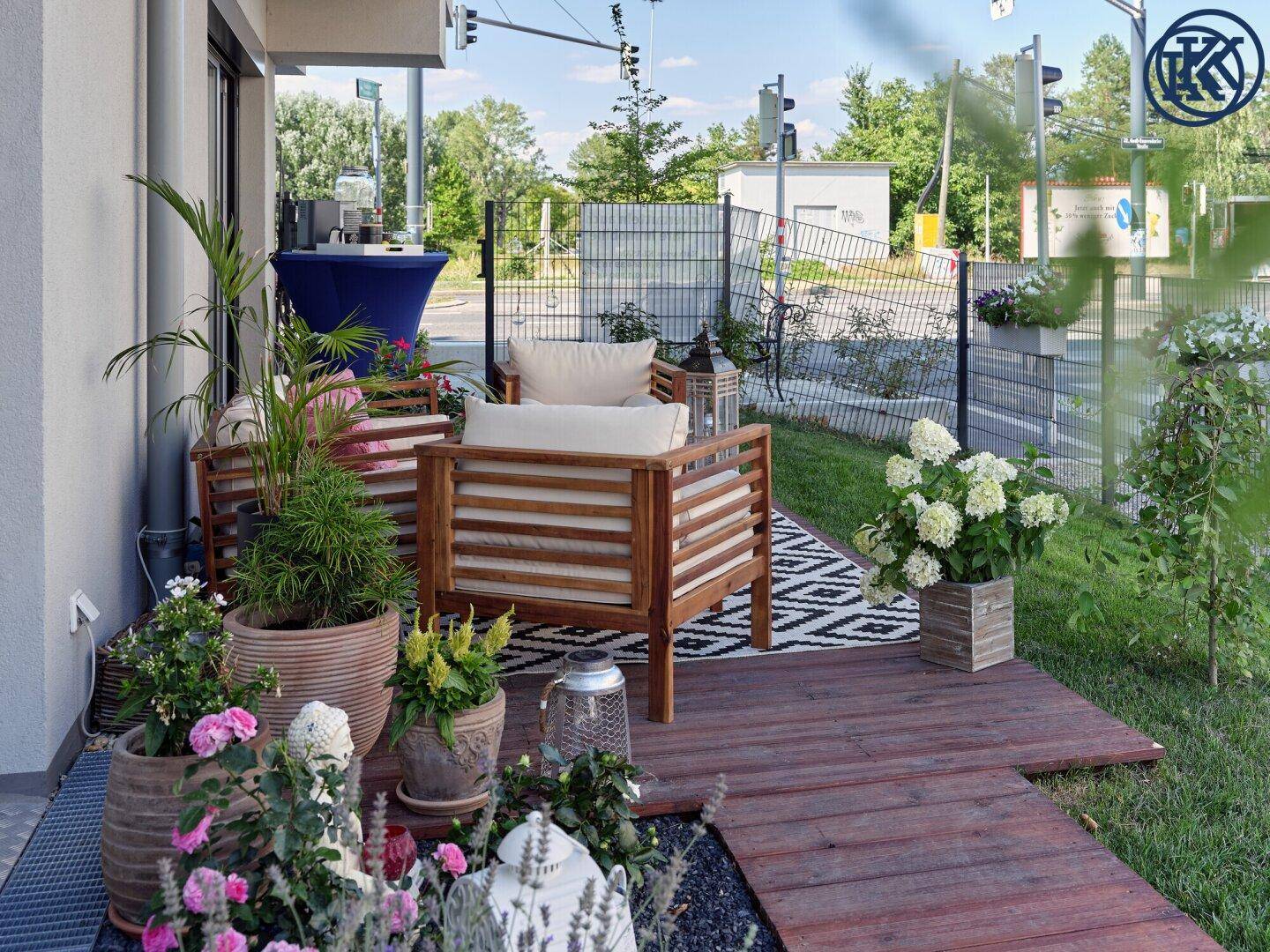Außenbereich mit Holzterrasse, Gartenmöbeln und üppiger Bepflanzung, umgeben von einem Zaun.