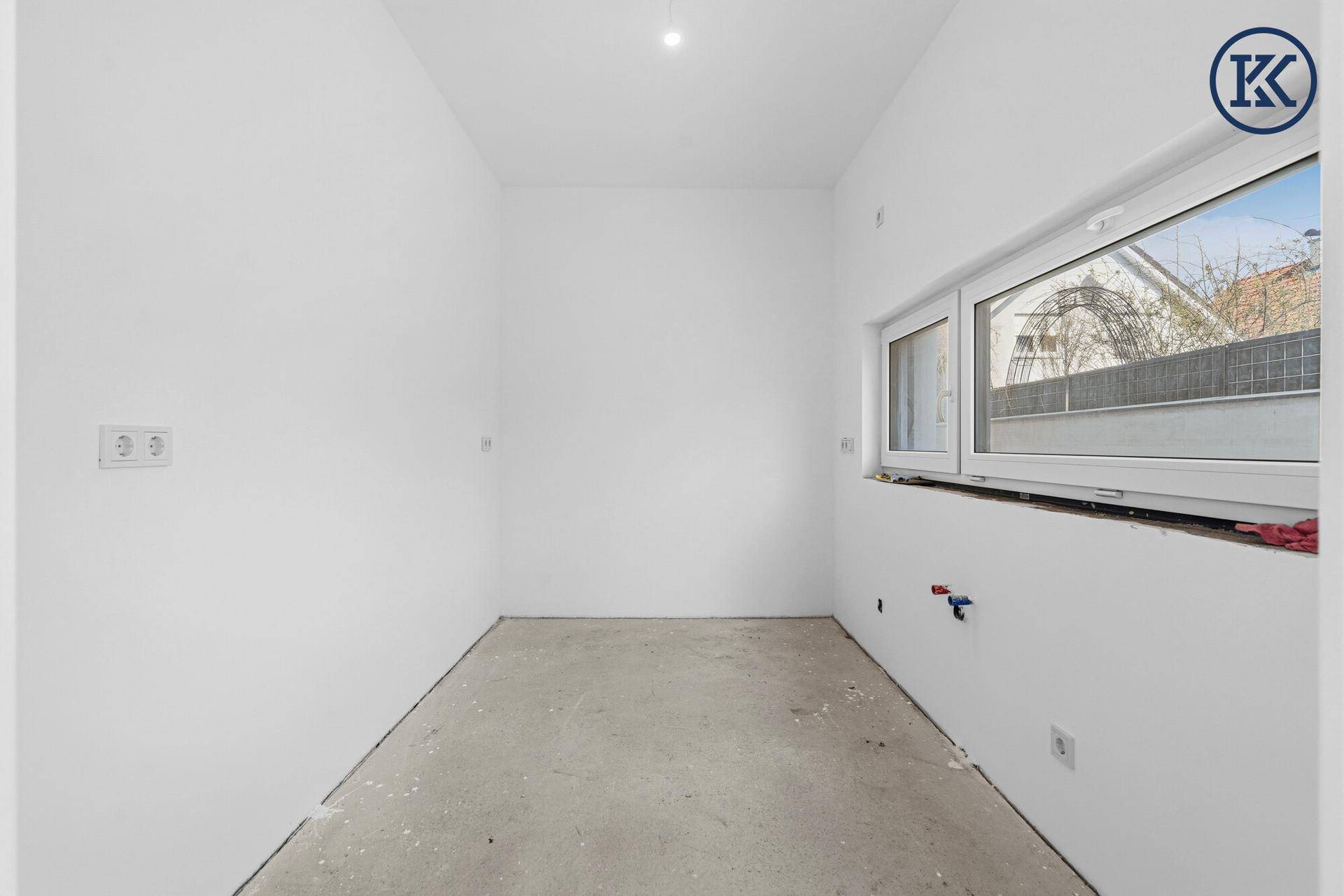 Unmöblierter Küchenraum im Rohbau mit Fenster und vorbereiteten Anschlüssen.