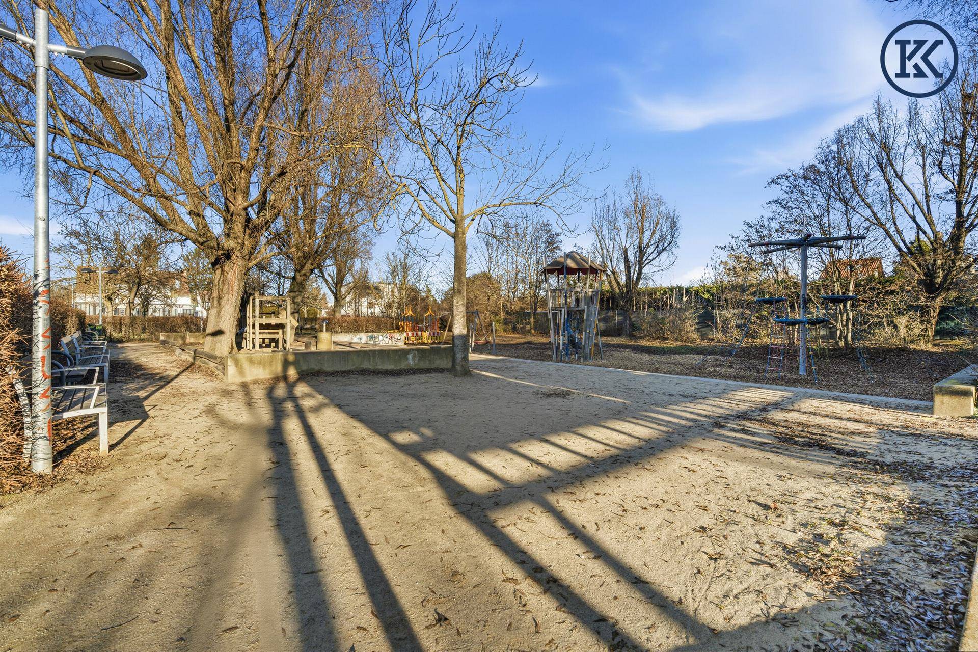 Öffentlicher Spielplatz mit Sandfläche, Bäumen und verschiedenen Spielgeräten.