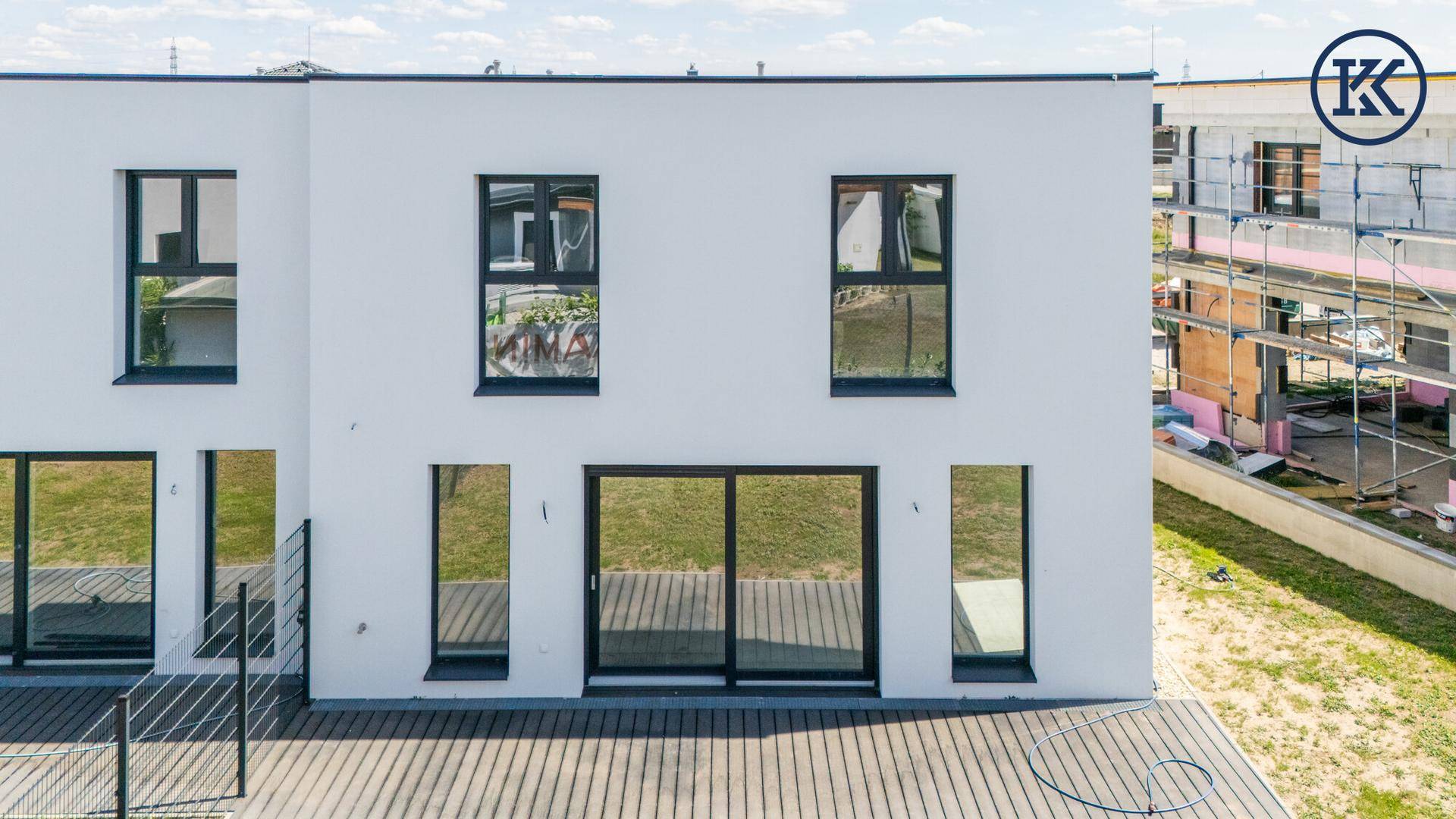 Rückseite des Neubaus mit großen Fenstern, Schiebetür und einer Holzterrasse, umgeben von Grünflächen.