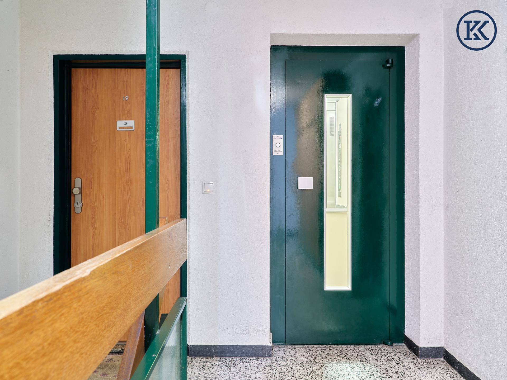 Flur im Gebäude mit zwei Türen und einem Aufzug, der auf die Etagen führt.