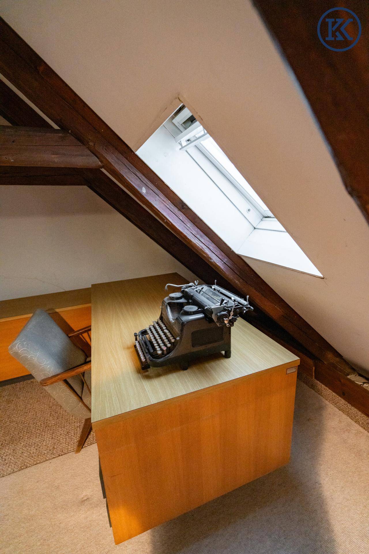 Schreibtisch mit alter Schreibmaschine im Dachgeschoss, beleuchtet durch ein Dachfenster.