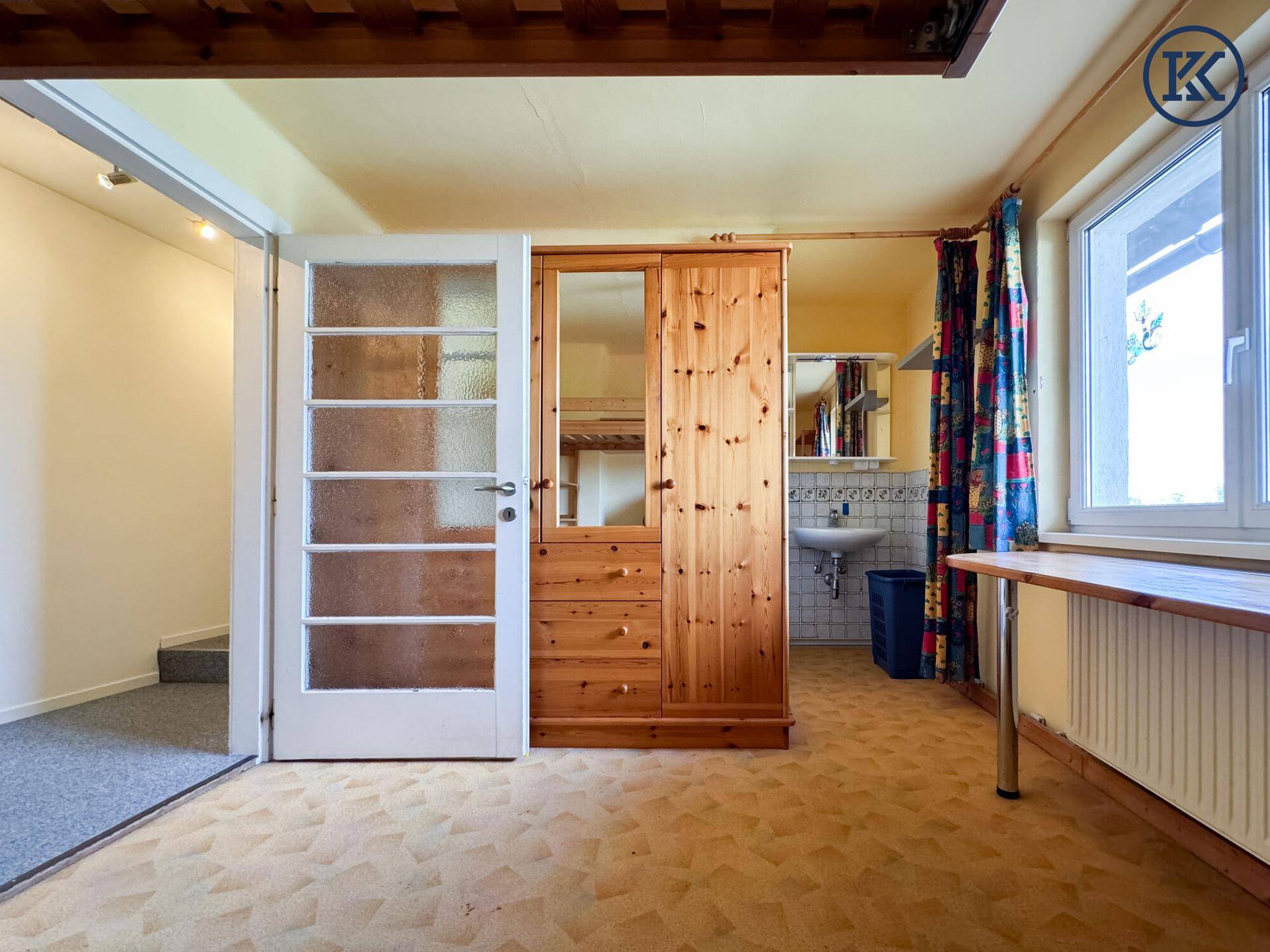 Schlafzimmer mit Holzschrank, Schreibtisch und einem kleinen Waschbeckenbereich in der Ecke.