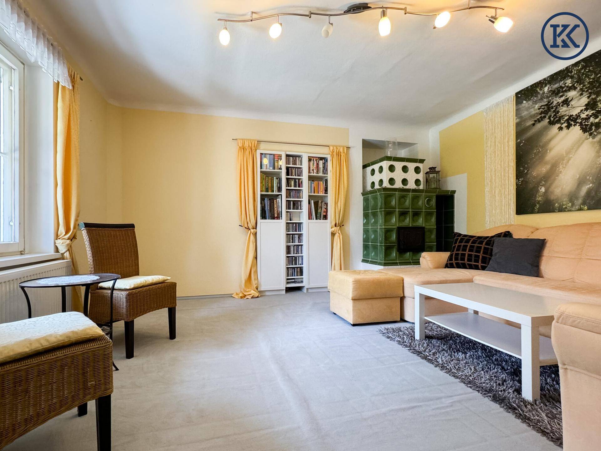 Helles Wohnzimmer mit Teppichboden, beigem Sofa und einem grünen Kachelofen, mit viel Tageslicht.