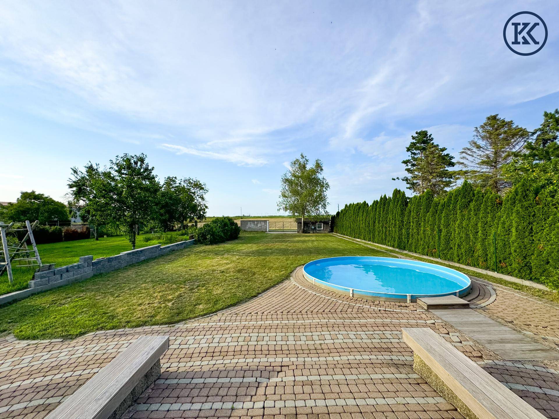 Weitläufiger Garten mit Pool, gepflastertem Bereich und Holzbänken unter blauem Himmel.