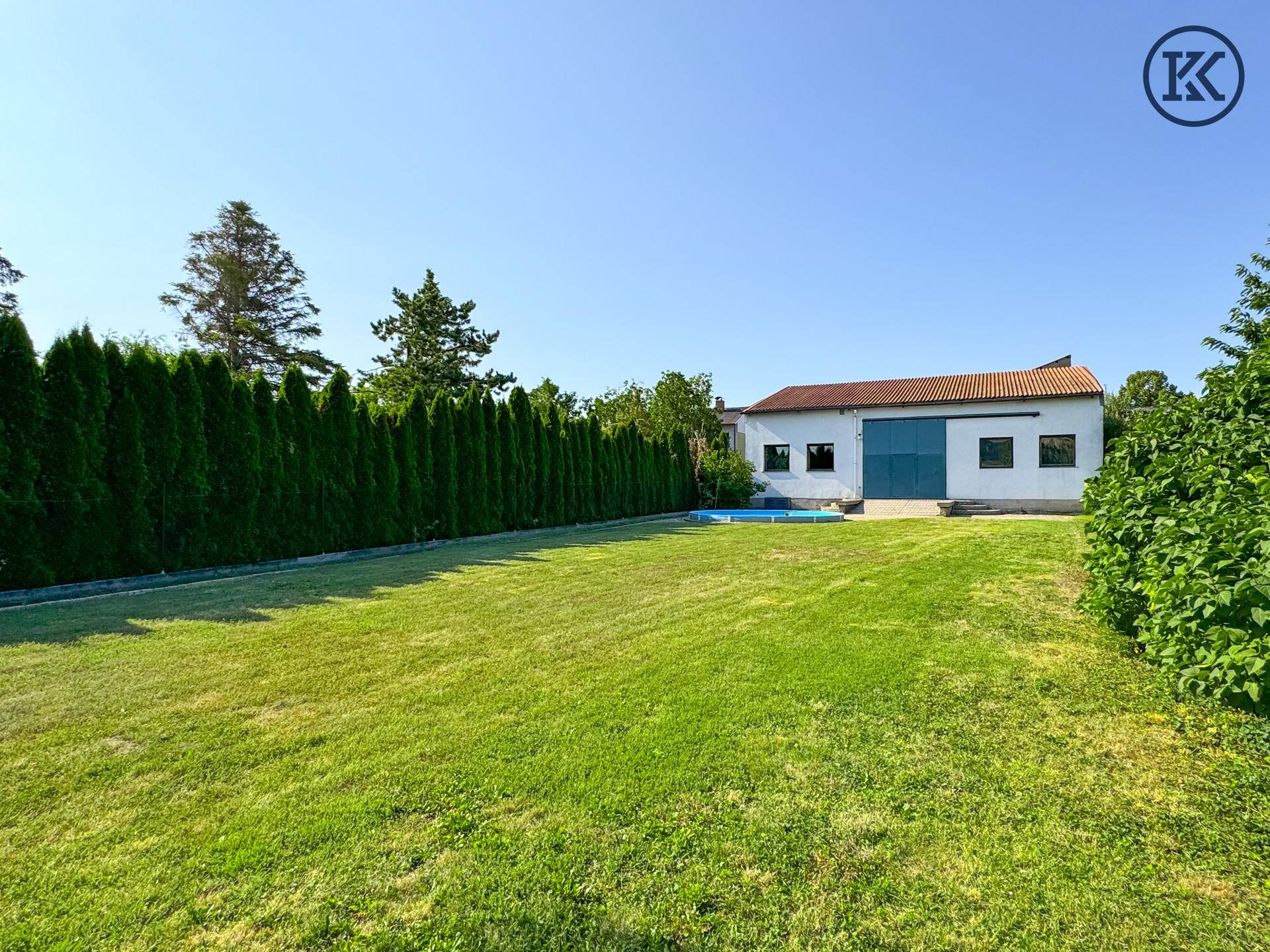 Weitläufiger grüner Garten mit hoher Hecke und Blick auf das Nebengebäude mit blauen Toren.