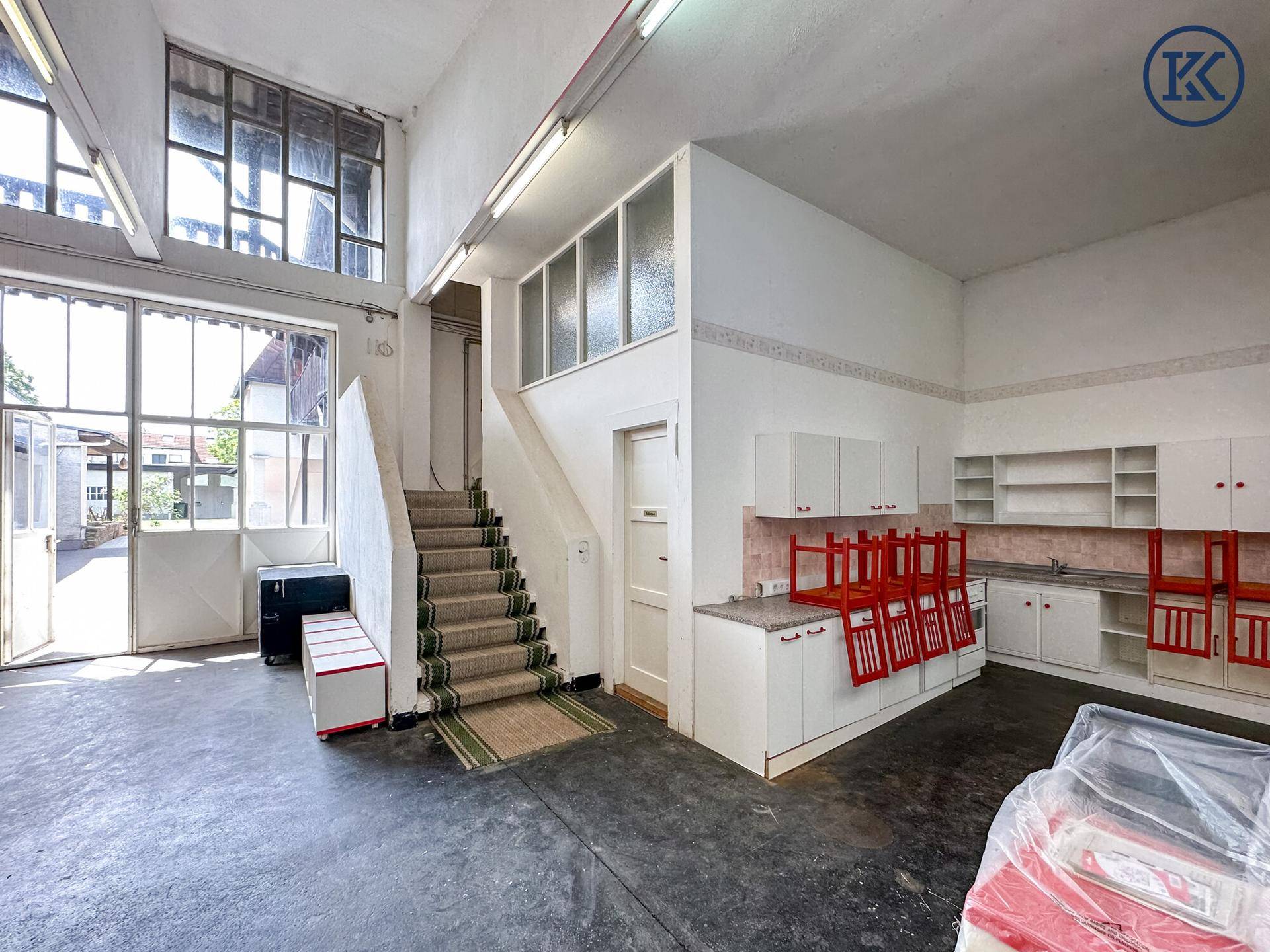 Großer offener Raum mit kleiner Küchenzeile, Treppe und hohen Fenstern, ideal als Werkstatt.