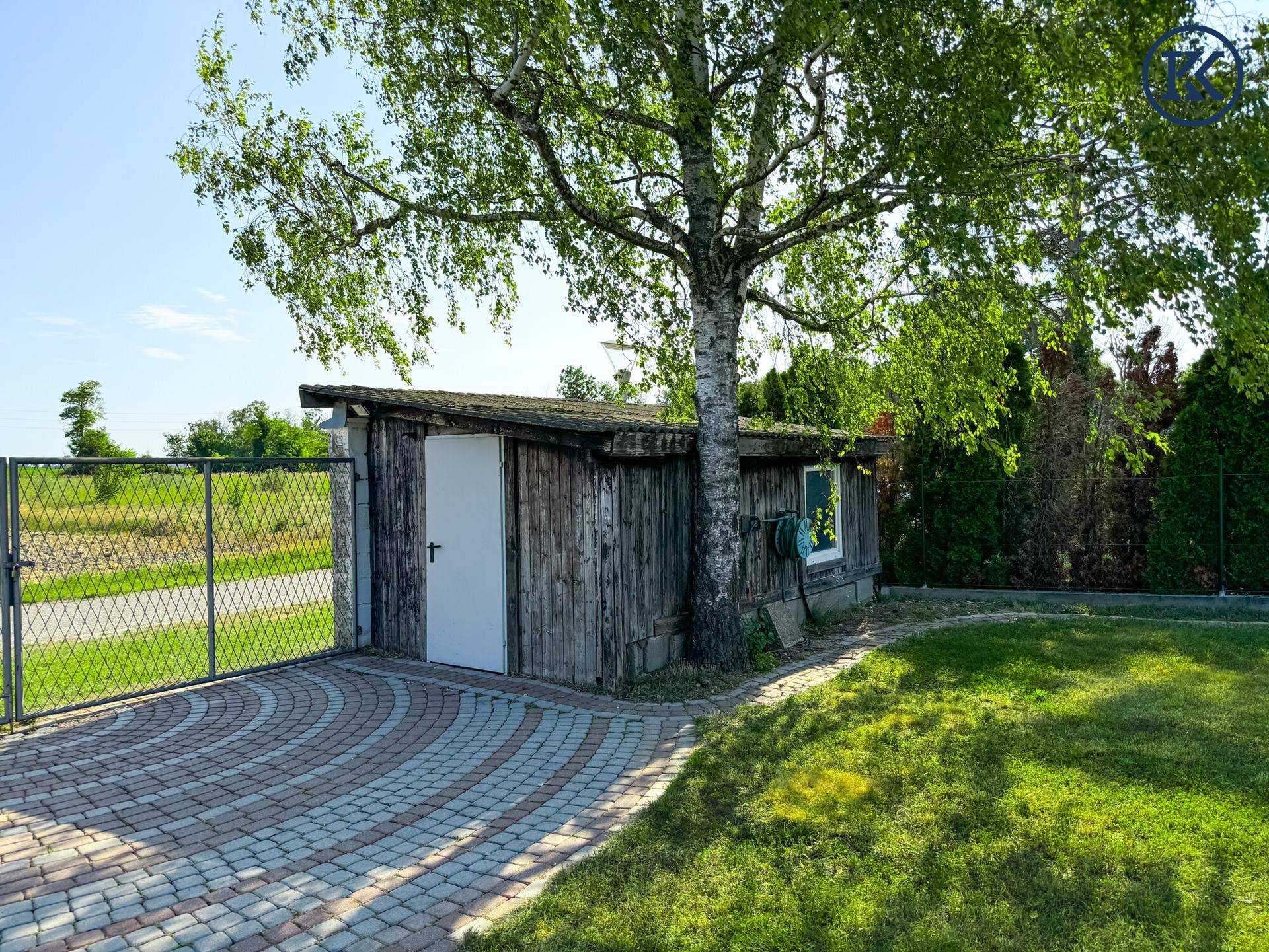 Rustikaler Holzschuppen neben einem gepflasterten Weg und einem Metalltor im Gartenbereich.
