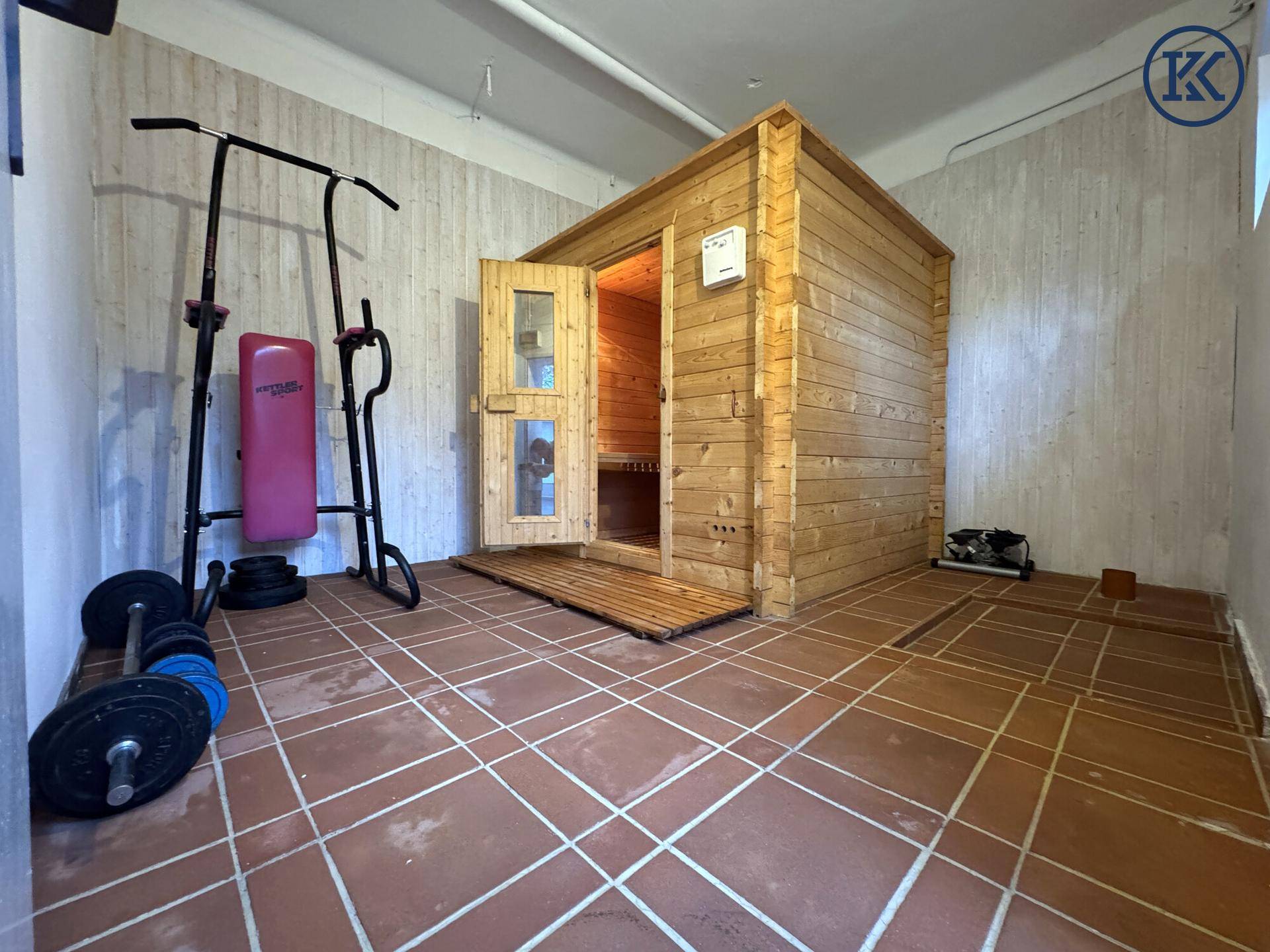 Fitnessraum mit einer freistehenden Sauna, Trainingsgeräten und Fliesenboden.